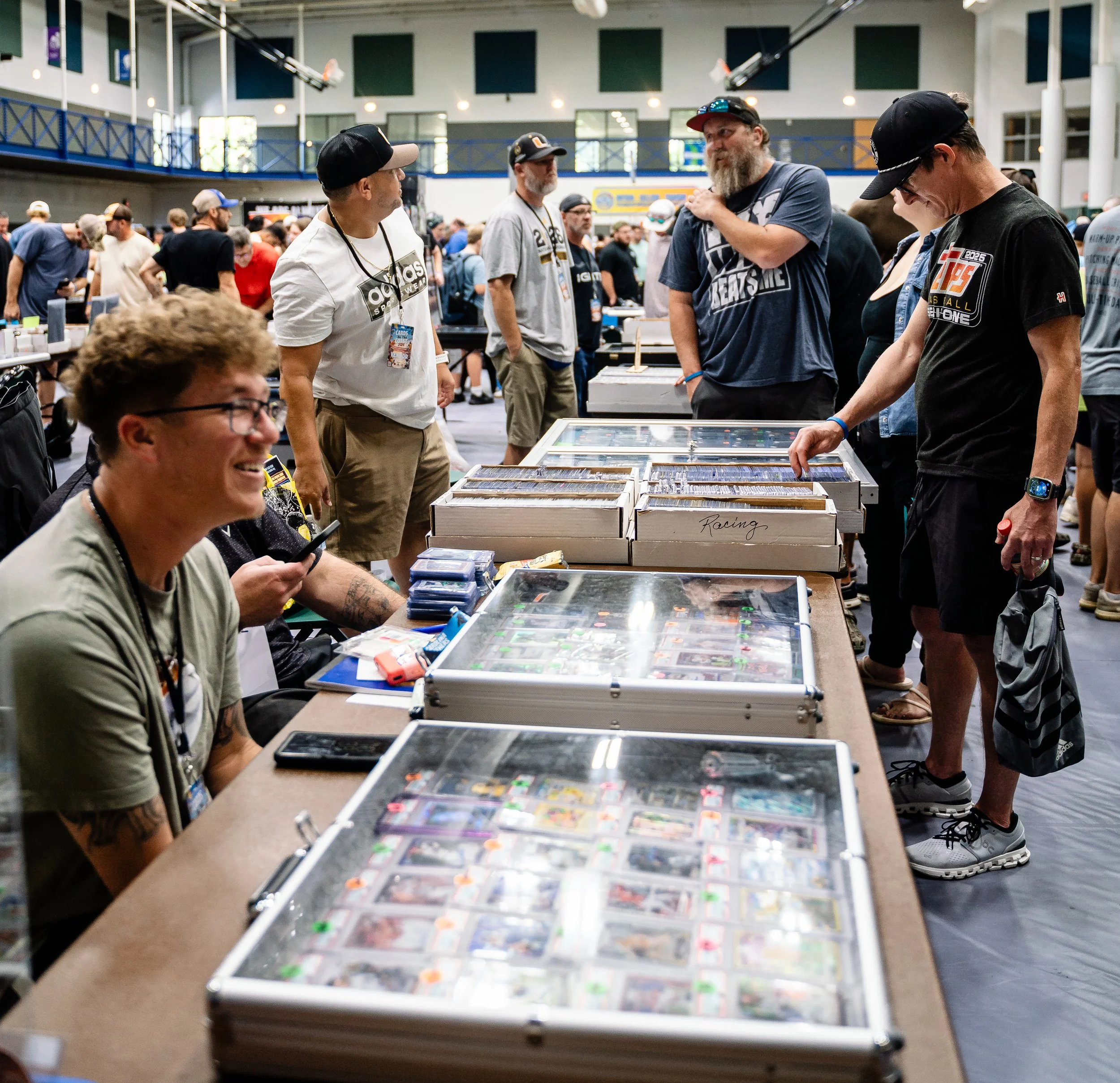 People browsing through collectible trading cards on display tables at a convention or event. The setting appears to be a large indoor space with multiple people engaged in exploration and discussion.
