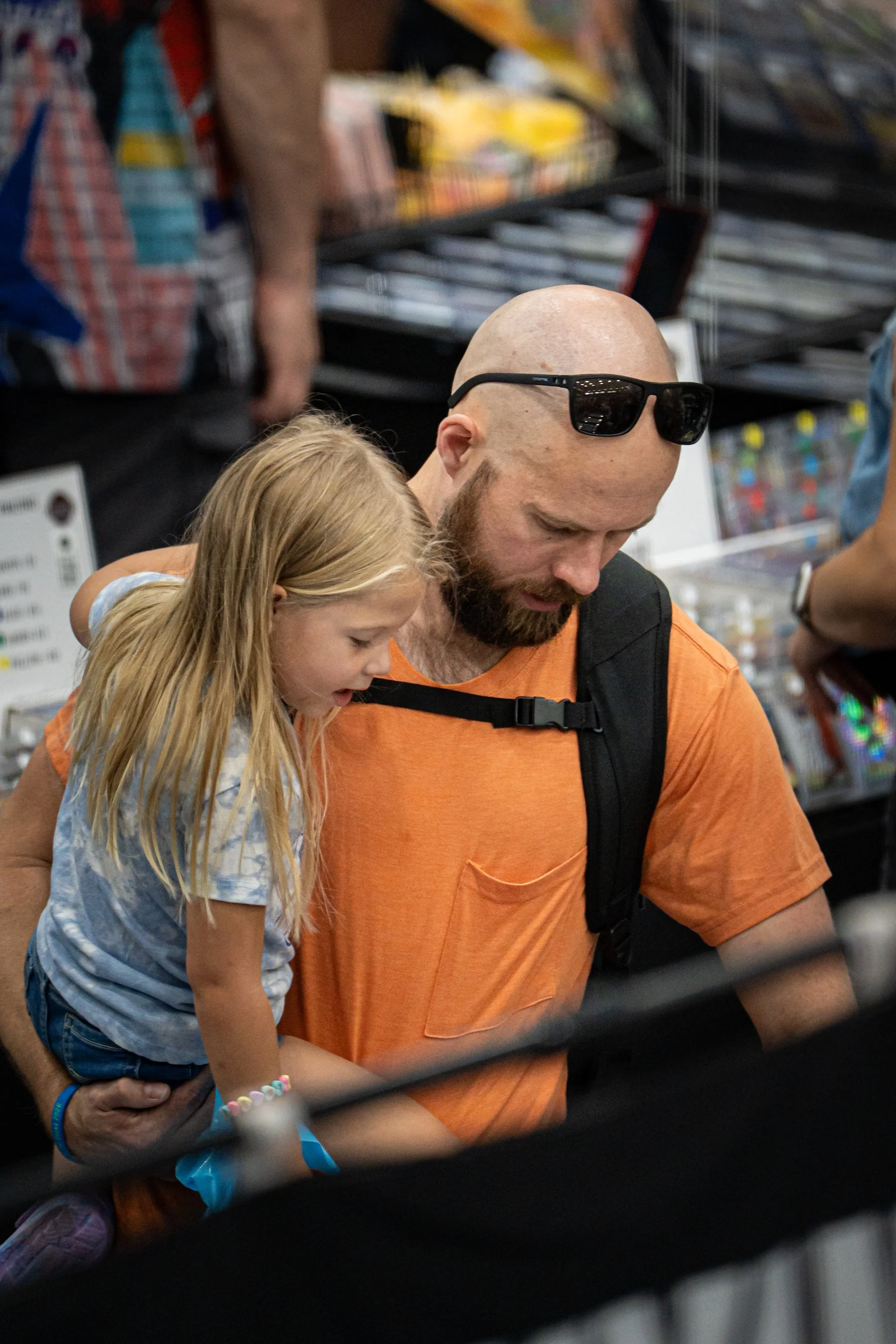 A man with a beard wearing sunglasses and an orange T-shirt is holding a young girl with blonde hair in a ponytail, wearing a light blue and white shirt and colorful bracelets, in a store aisle with shelves of products in the background.