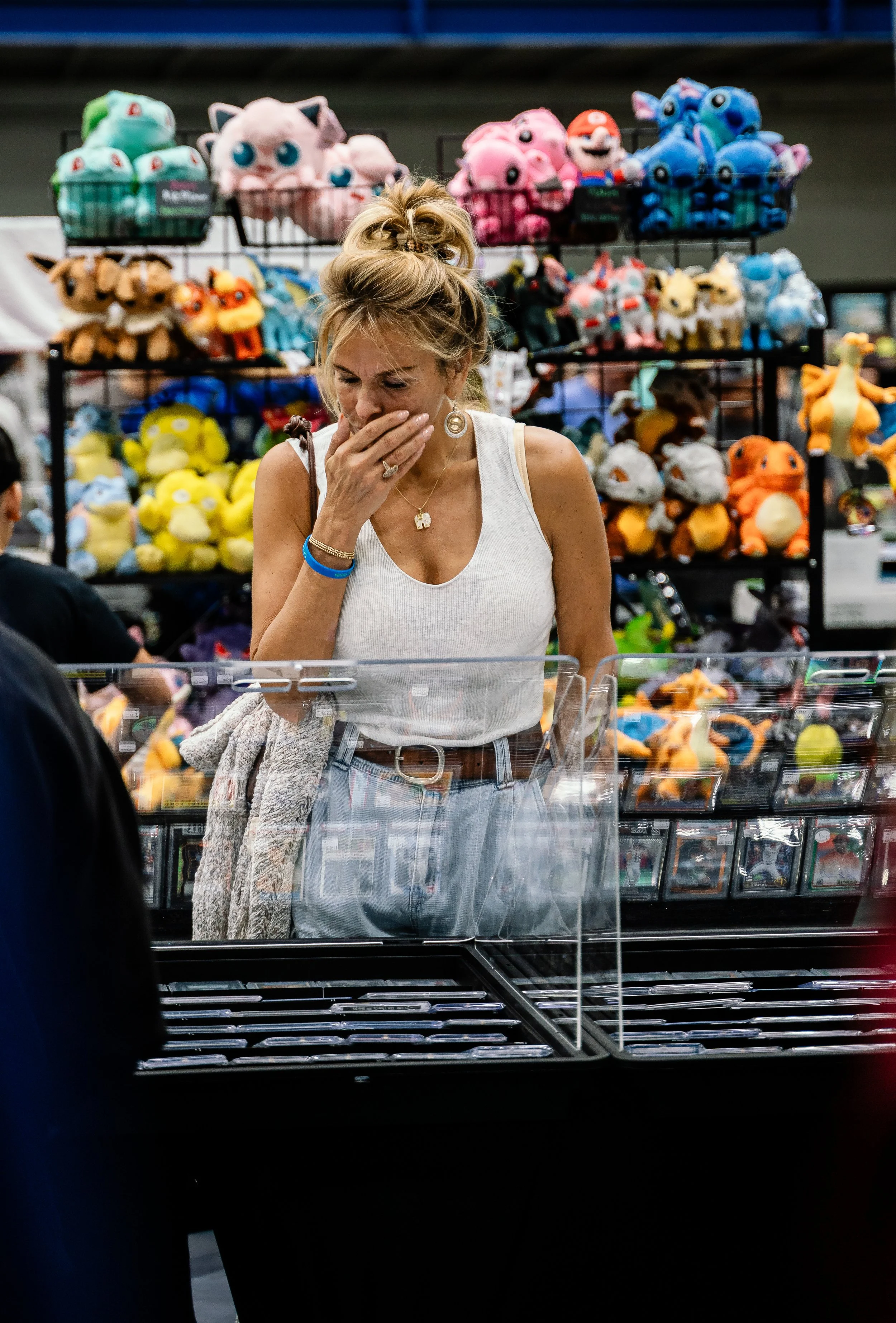 A woman wearing a white tank top and jewelry is shopping at a toy booth at an indoor event. She appears to be surprised or confused, with her hand covering her mouth. The booth has shelves filled with plush toys of various characters, including Pokém