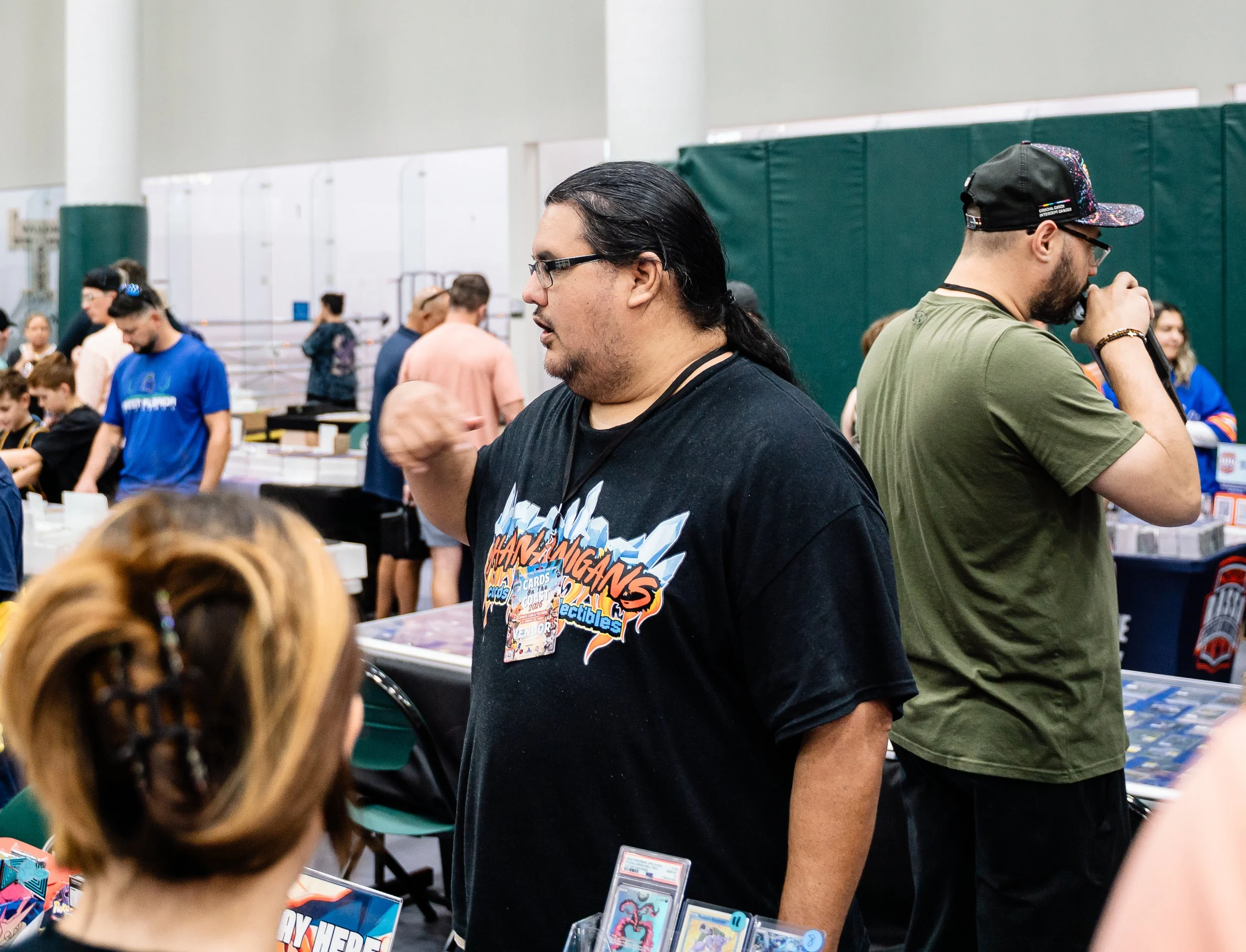 People at a tabletop gaming event, playing card games, with spectators behind them in a gymnasium.