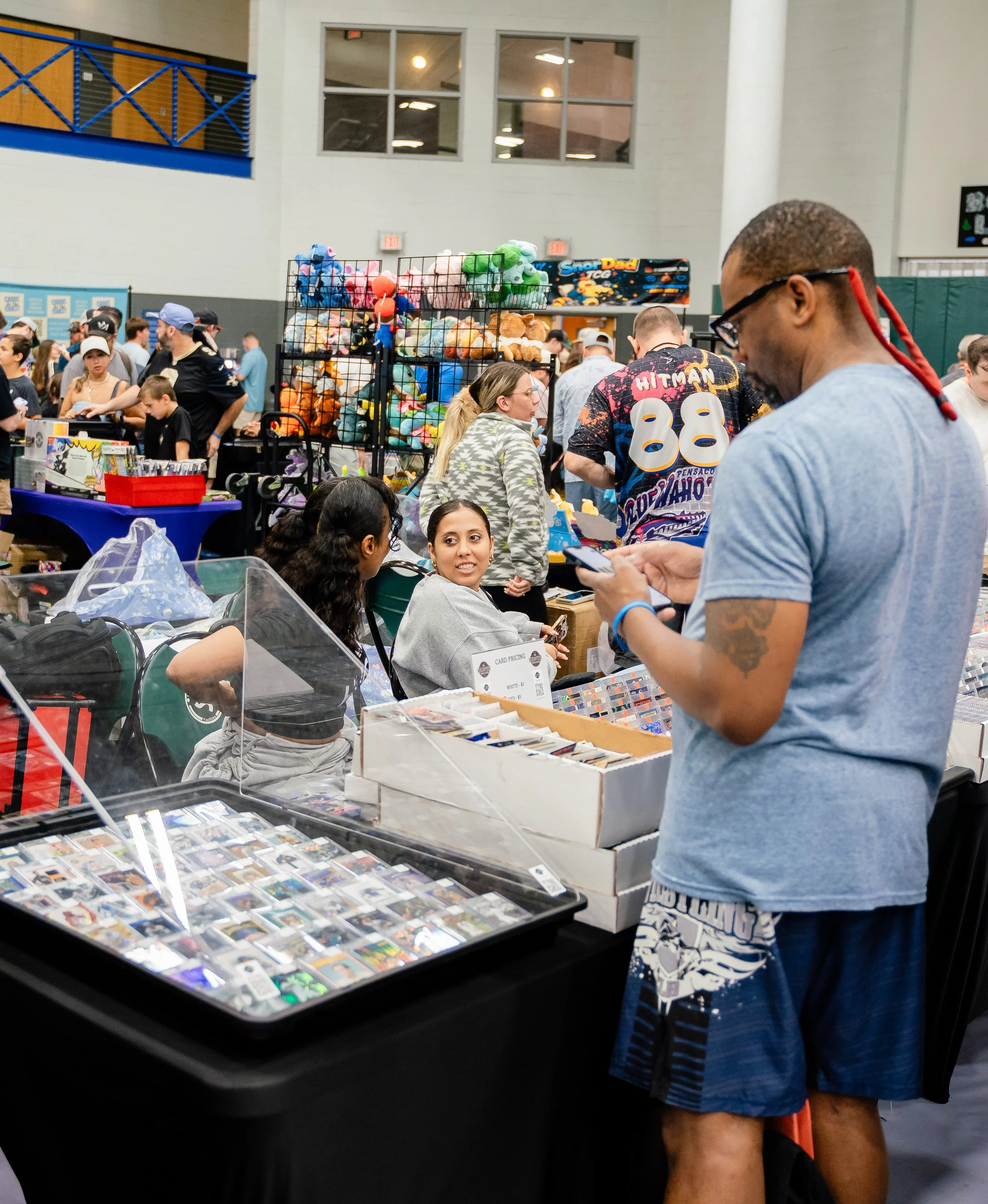 A man with glasses and a tattooed arm is looking at a display of trading cards or collectible cards at an indoor event. The display contains numerous cards with colorful images. There are other people in the background, and a rack of plush toys is vi