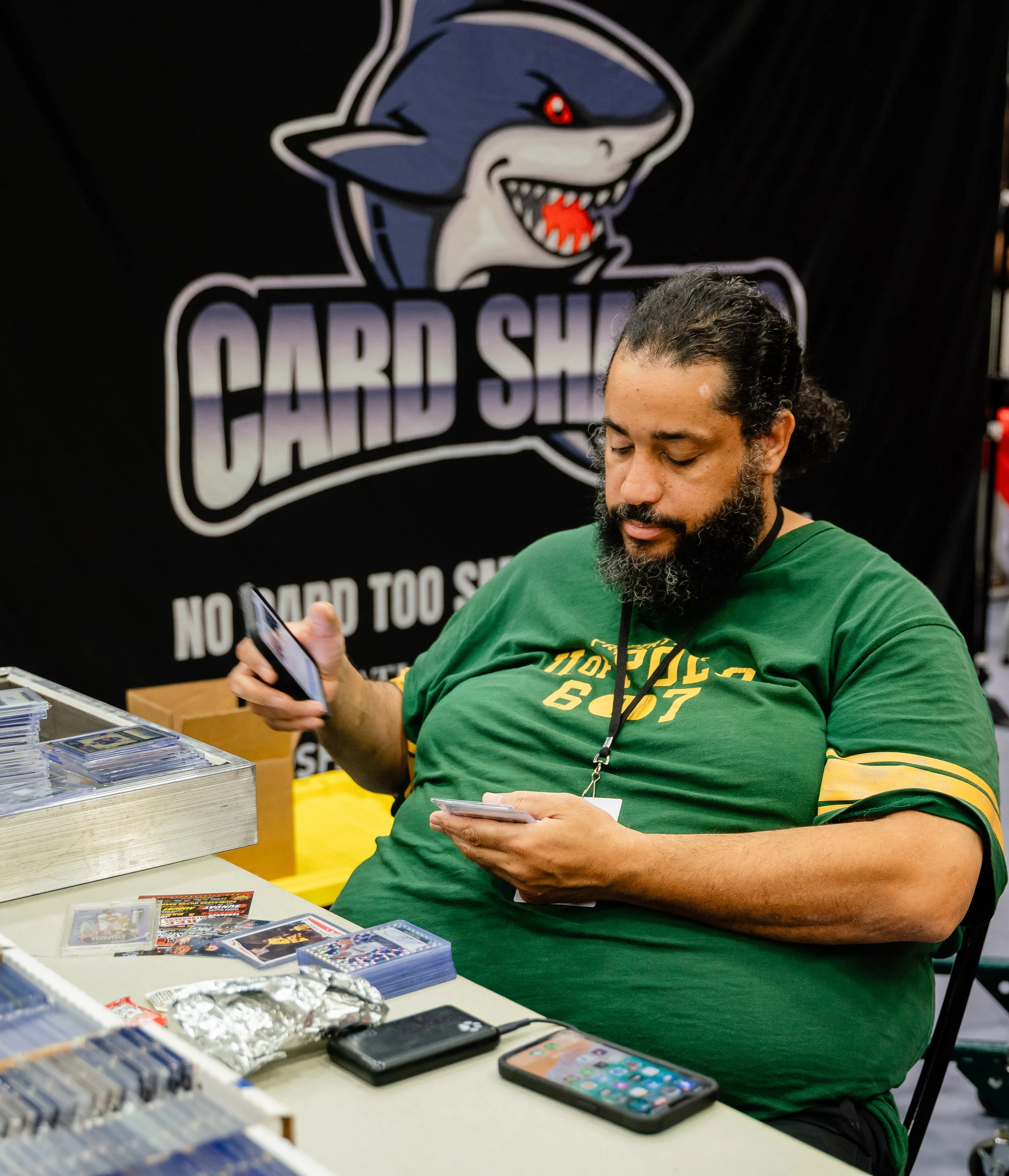 A man with a beard and curly hair sitting at a table looking at his phone and surrounded by various trading cards and trading card packs, with a large logo of a cartoon shark with glowing red eyes and sharp teeth on a black background behind him.