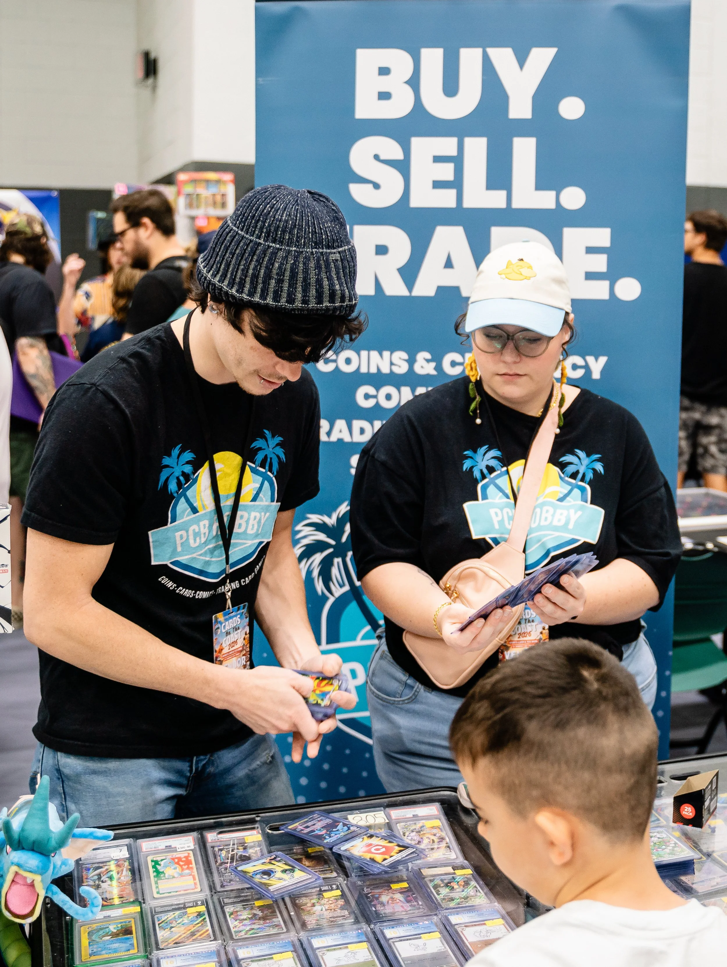 Two people browsing trading cards at an event, with a large blue sign behind them that says "BUY. SELL. TRADE."