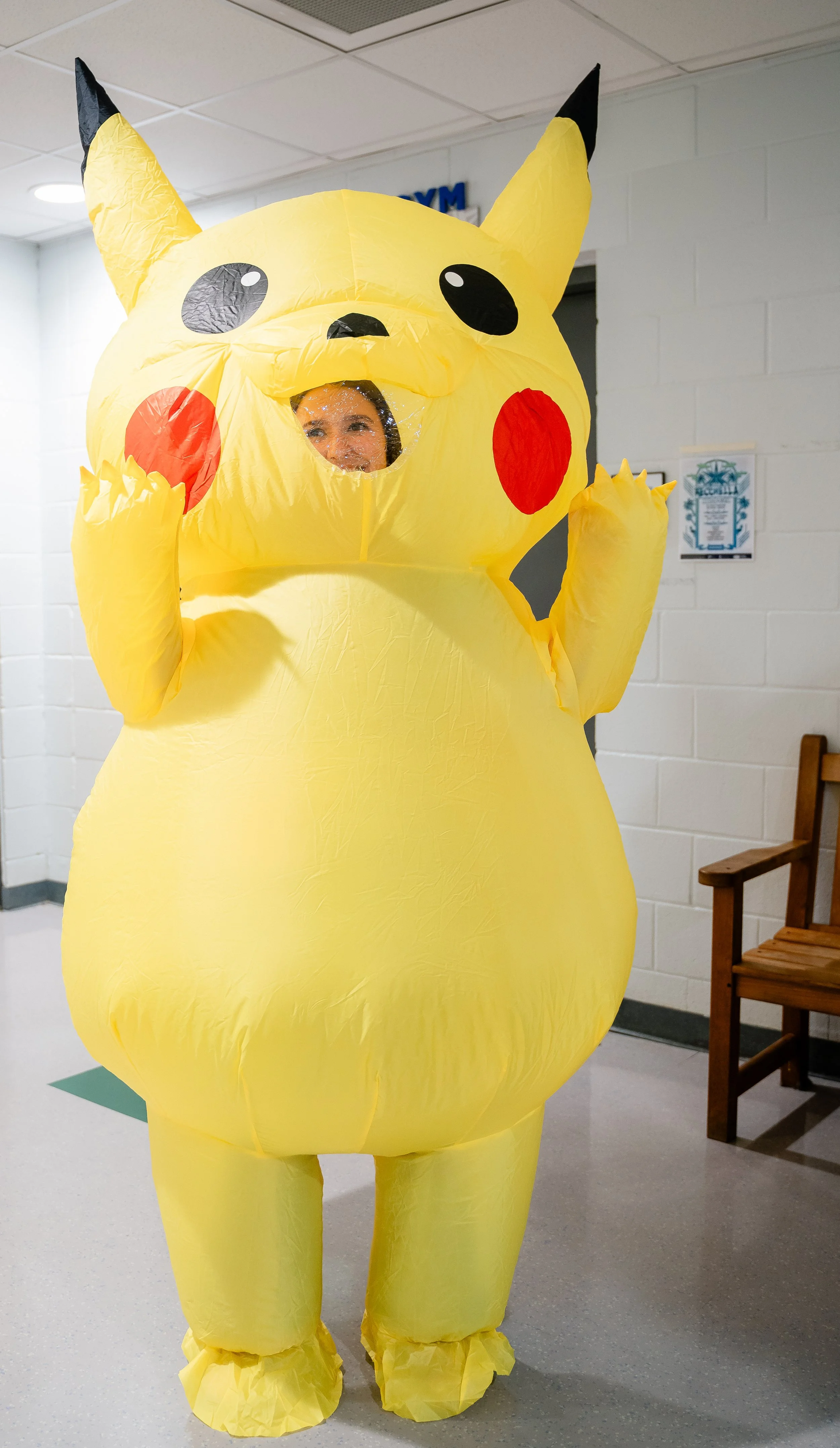 Person dressed in a Pikachu costume with a large Pikachu head, standing indoors near a white tile wall and wooden chairs, showing the person's face through an opening in the face of the costume.