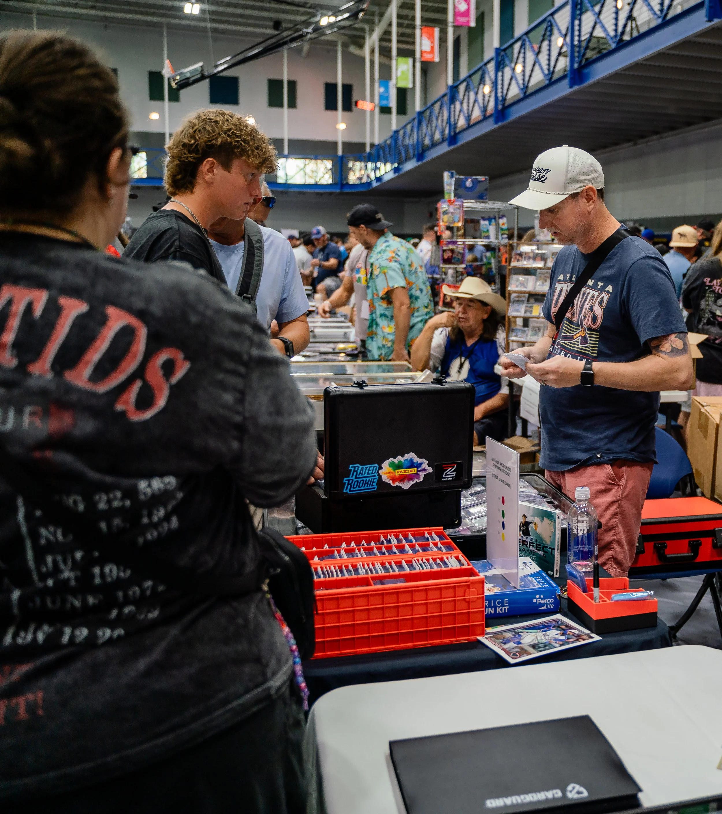 Inside a busy convention or trade show with people browsing displays of trading cards, collectibles, and memorabilia. A man in a blue Atlanta Falcons t-shirt and gray cap is examining items while others look on or browse nearby booths. The environmen