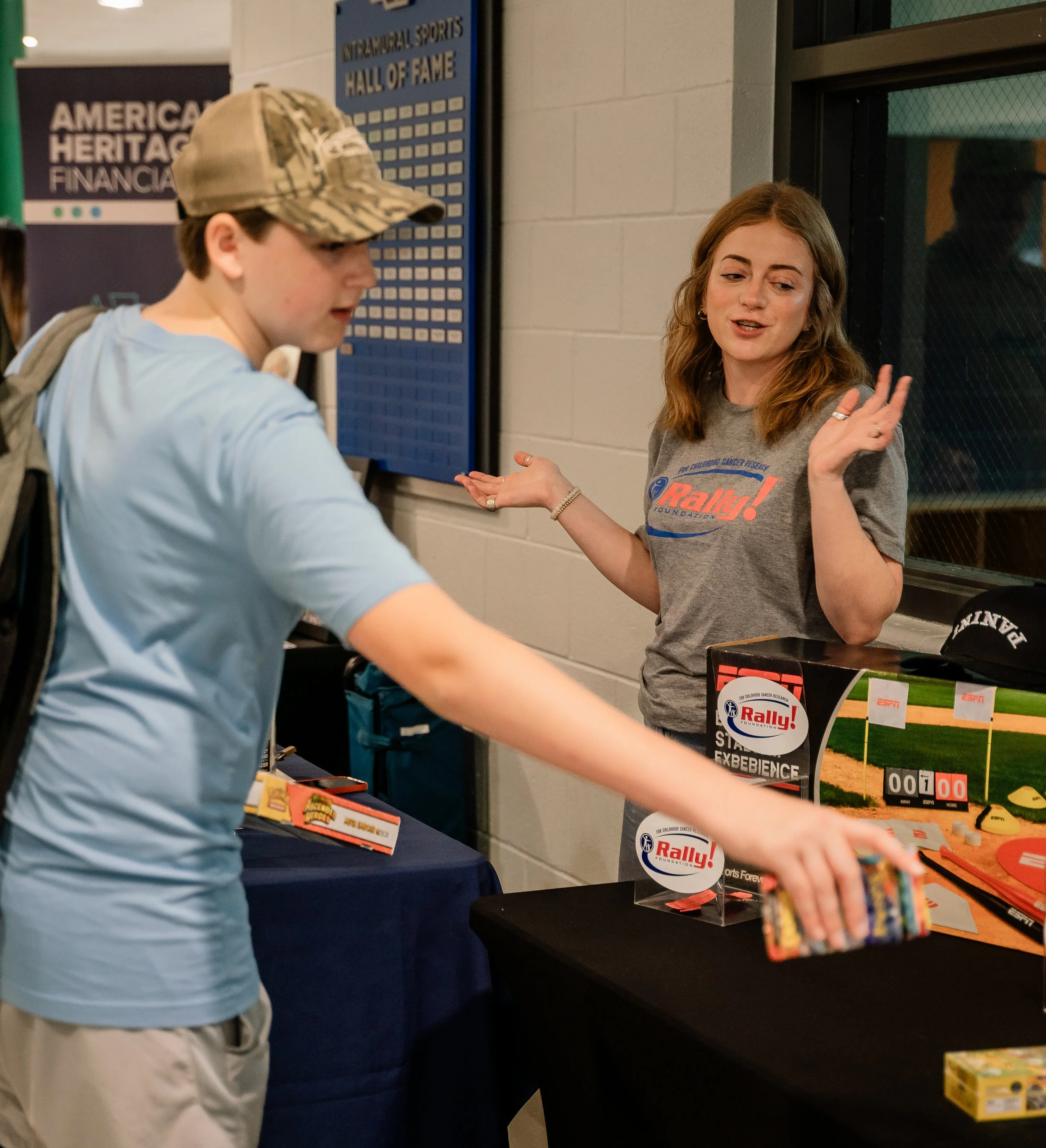 A woman with brown hair speaking and gesturing with her right hand, wearing a gray shirt with a sports foundation logo, standing near a table with brochures and promotional materials for the Rally Foundation and childhood cancer research. Behind her,