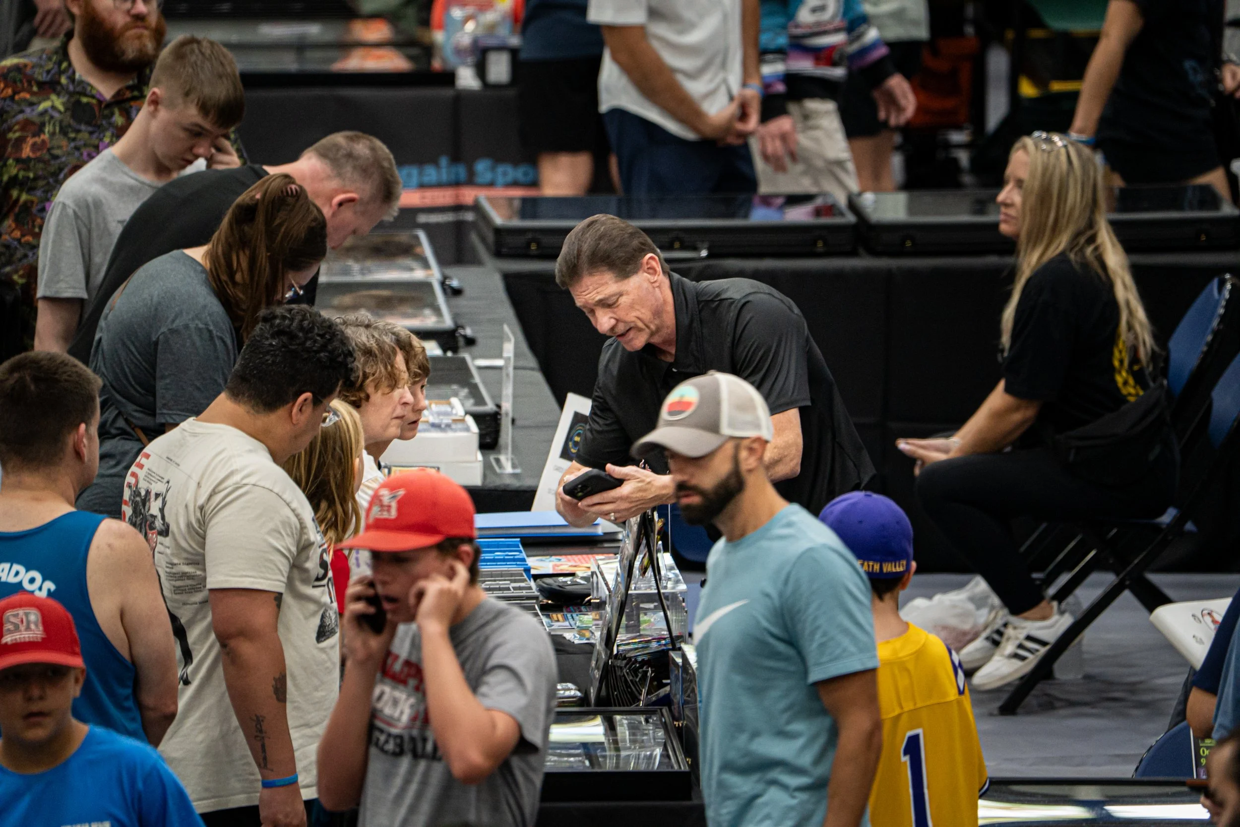 People browsing and purchasing items at a convention or expo, with various tables and displays and some individuals wearing sports jerseys and hats.