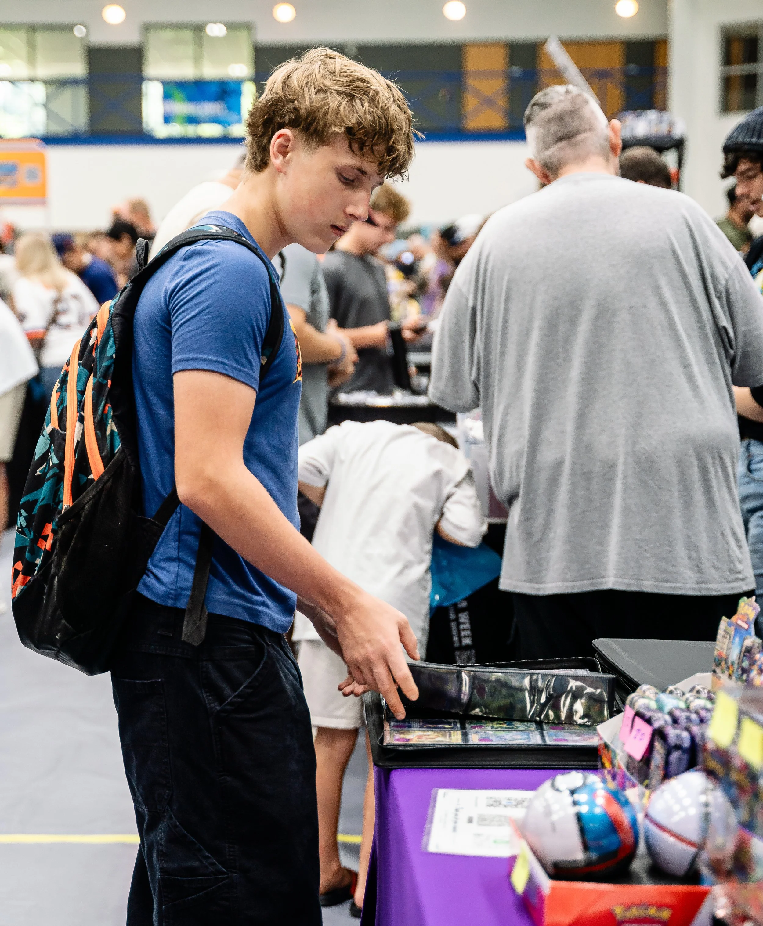 Young man with curly brown hair wearing a blue t-shirt and carrying a backpack shopping at a vendor table at an indoor market or event.
