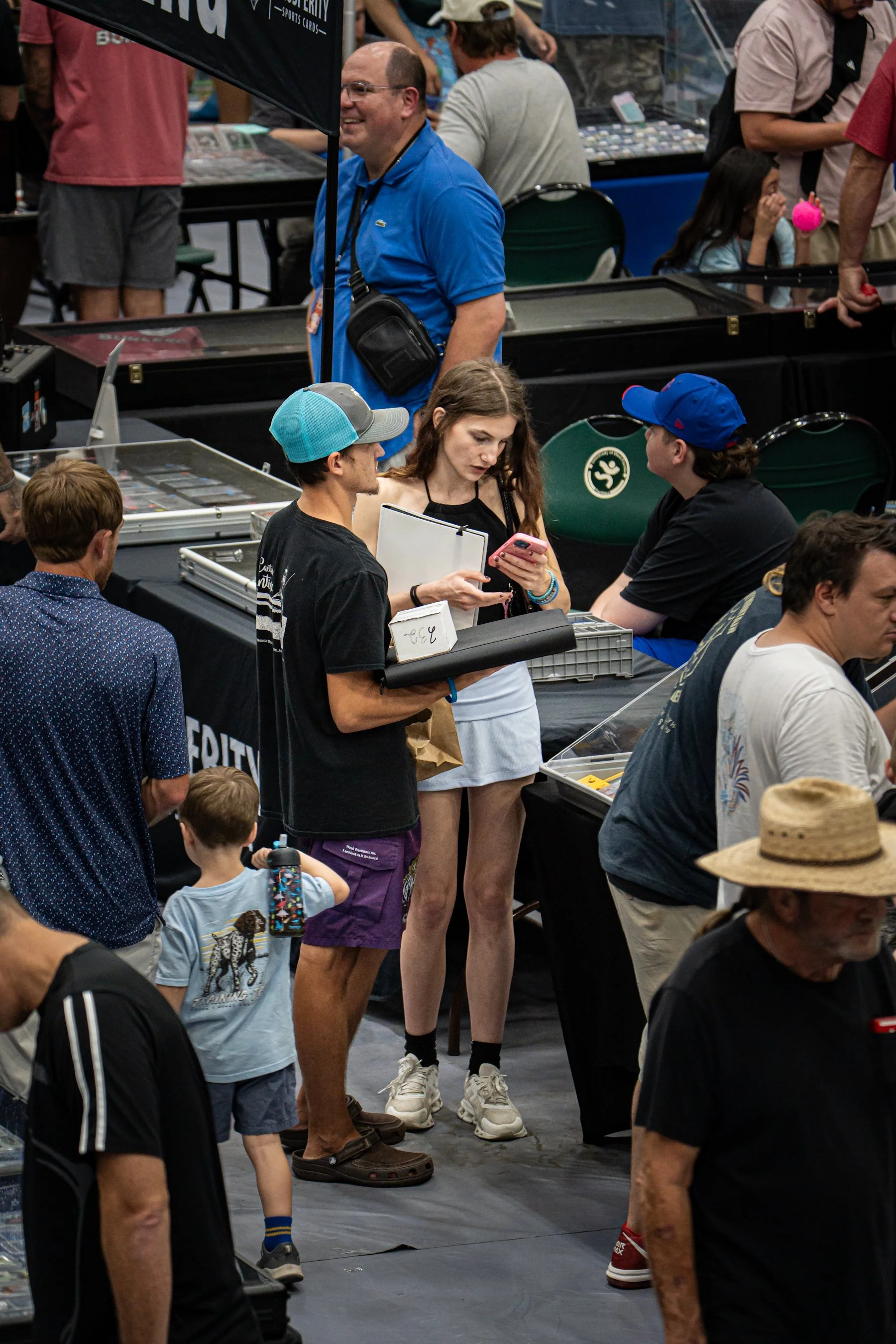 People browsing items at a convention or trade show, with most individuals standing and some sitting, surrounded by display booths and tables with collectibles or merchandise.