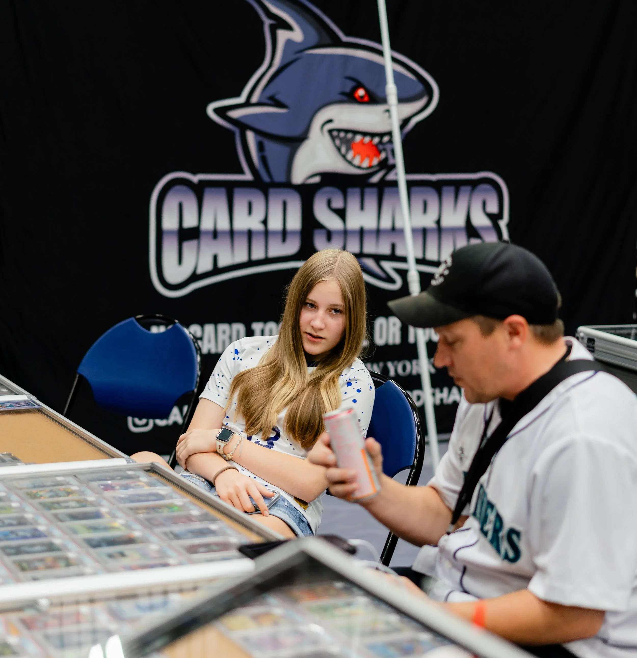 A young girl with long light brown hair sitting at a table with a woman, both at a card or trading game event. The woman is wearing a white shirt with blue spots and a smartwatch. The girl is looking towards the camera with a neutral expression. In t