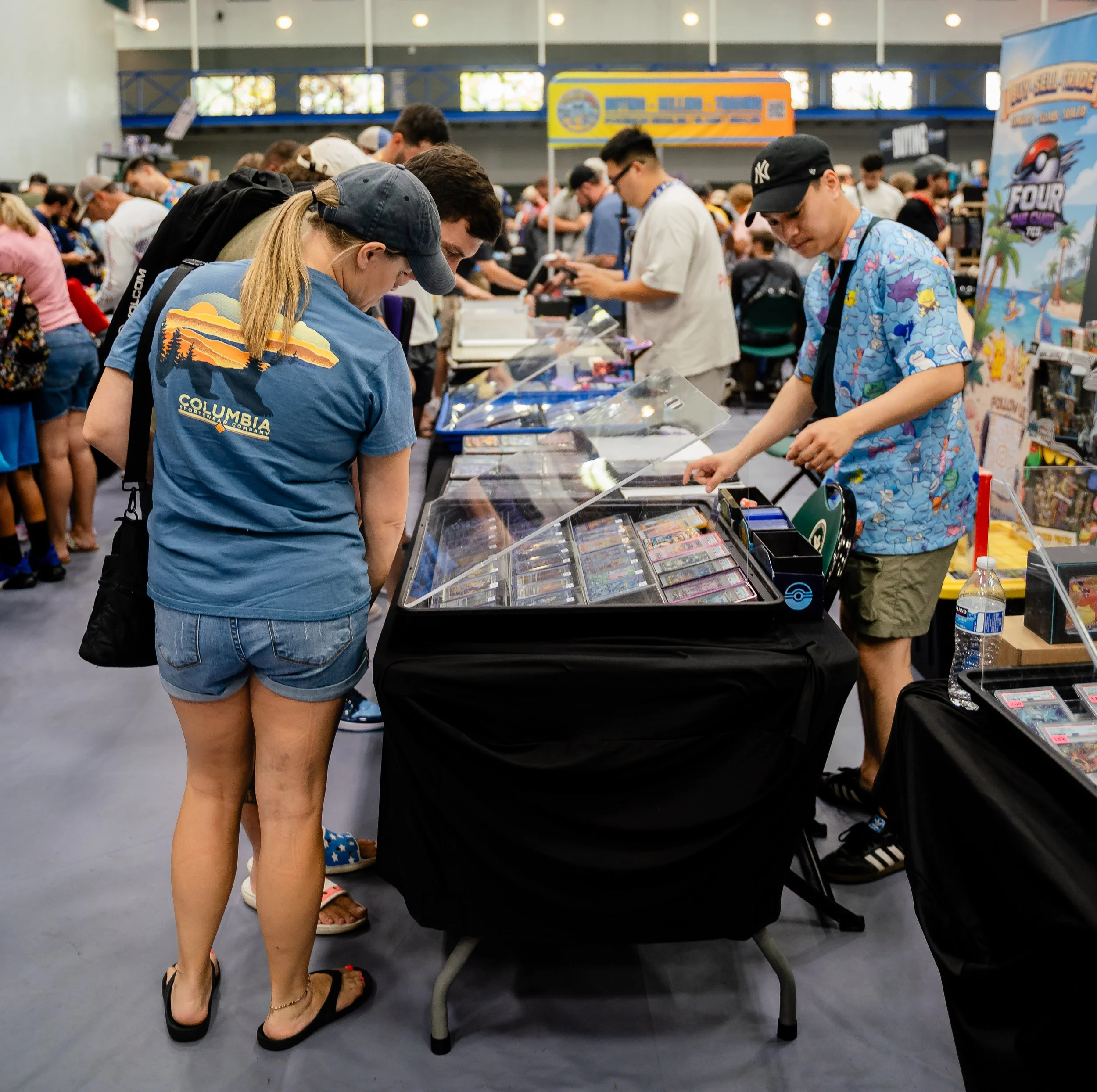 At a convention, people are browsing merchandise displayed on tables and shelves. Crowds of attendees are looking at items, some standing and others leaning over the display cases. The background shows signs and posters related to the event.