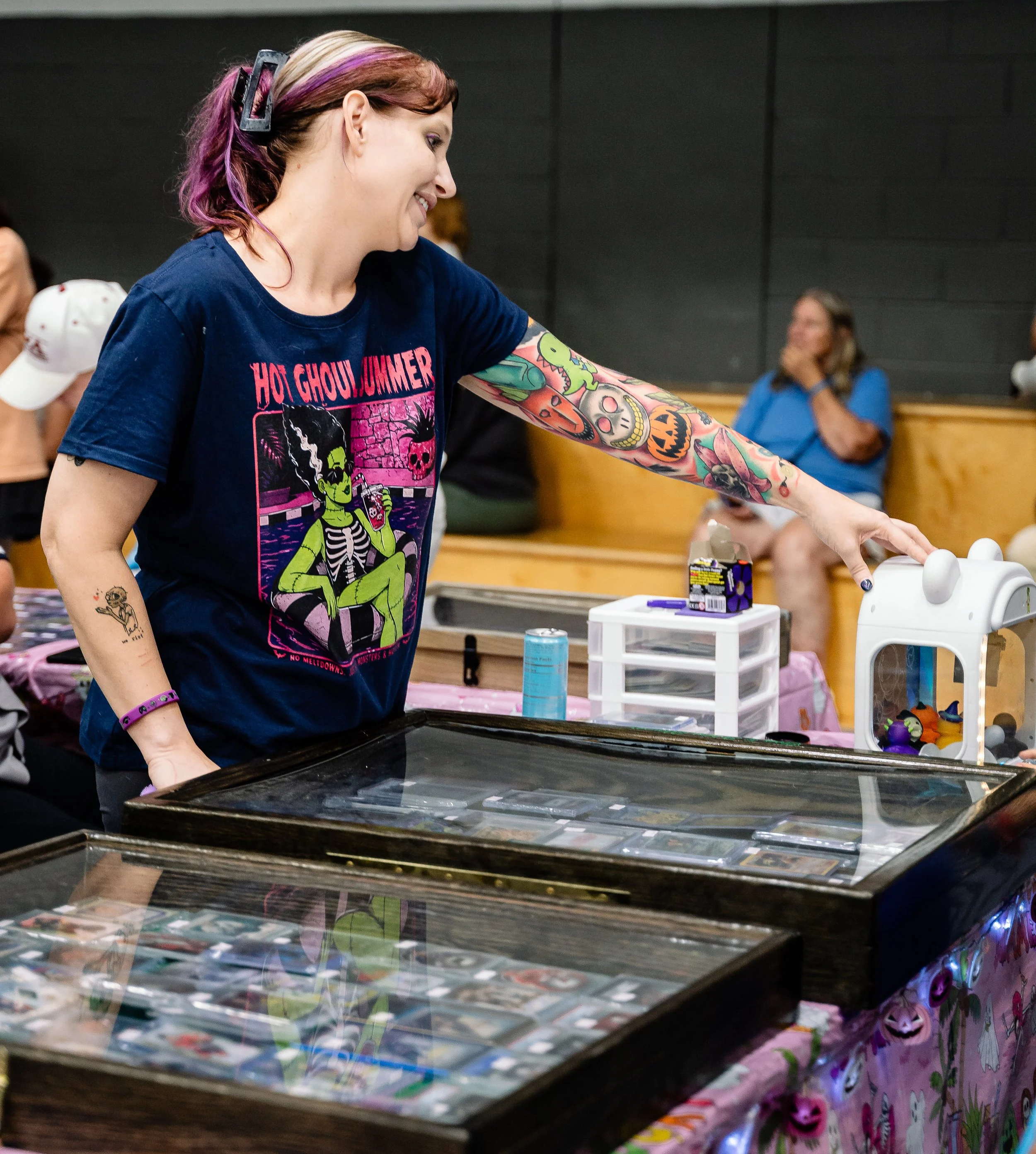 A woman with multicolored hair and colorful tattoos Smiling as she interacts with items on a table, in a room with dark walls and other people in the background.
