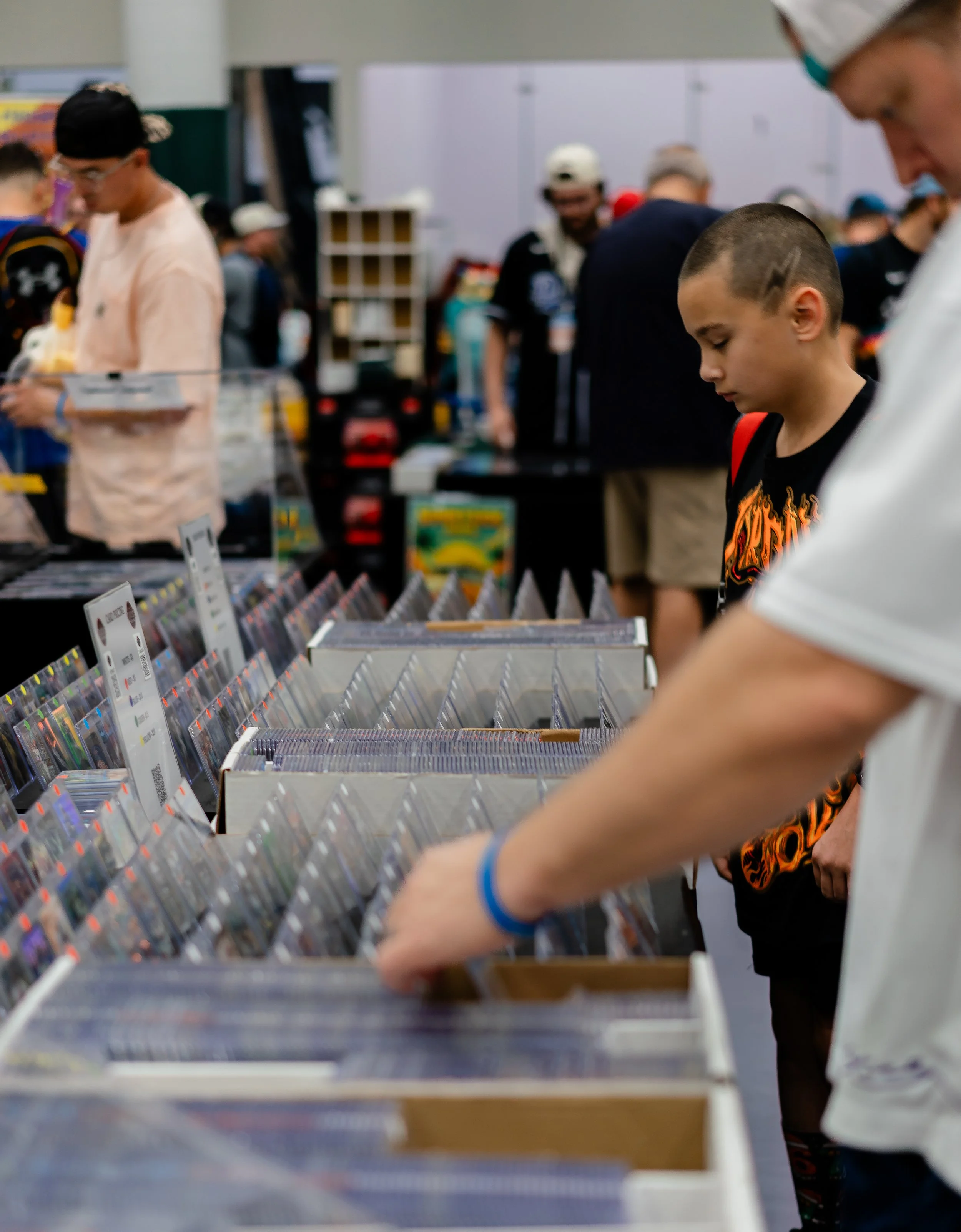 Several people browsing through a collection of comic books or trading cards at an indoor convention or store, with focus on a young boy with a buzz cut and a graphic T-shirt, and some adults nearby.