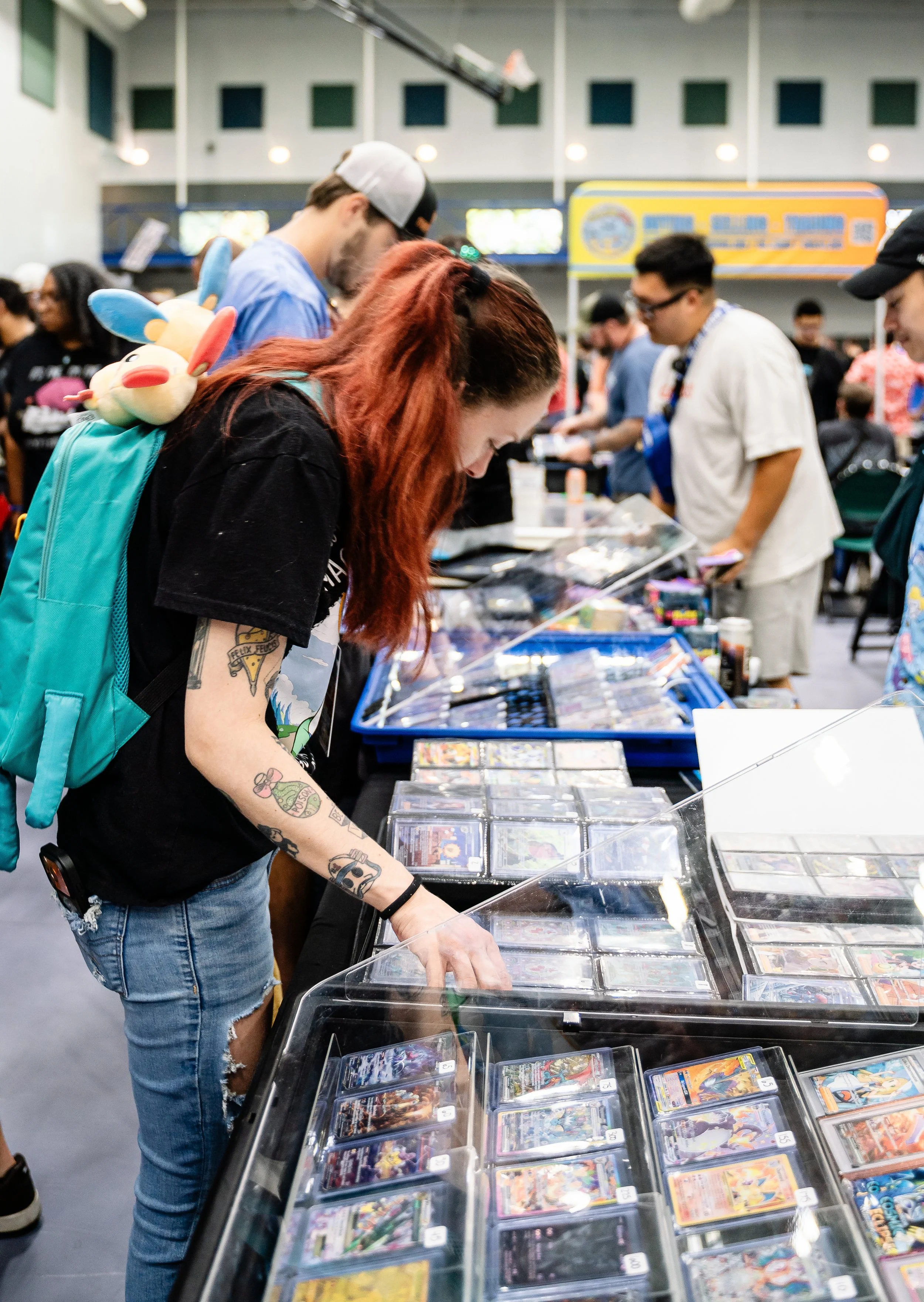 A young woman with red hair and tattoos on her arms looking at trading cards in a display case at a card shop or convention. She is wearing a black shirt and denim shorts, and has a blue backpack with a plush toy attached to it. Other people browse s