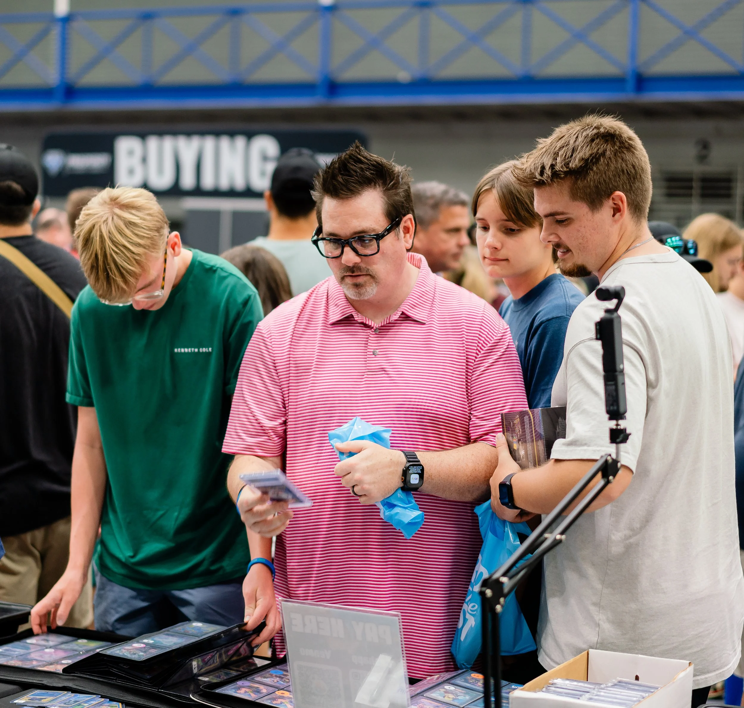 People shopping at a market stall with trading cards, with a sign in the background that says 'BUYING' and other shoppers.