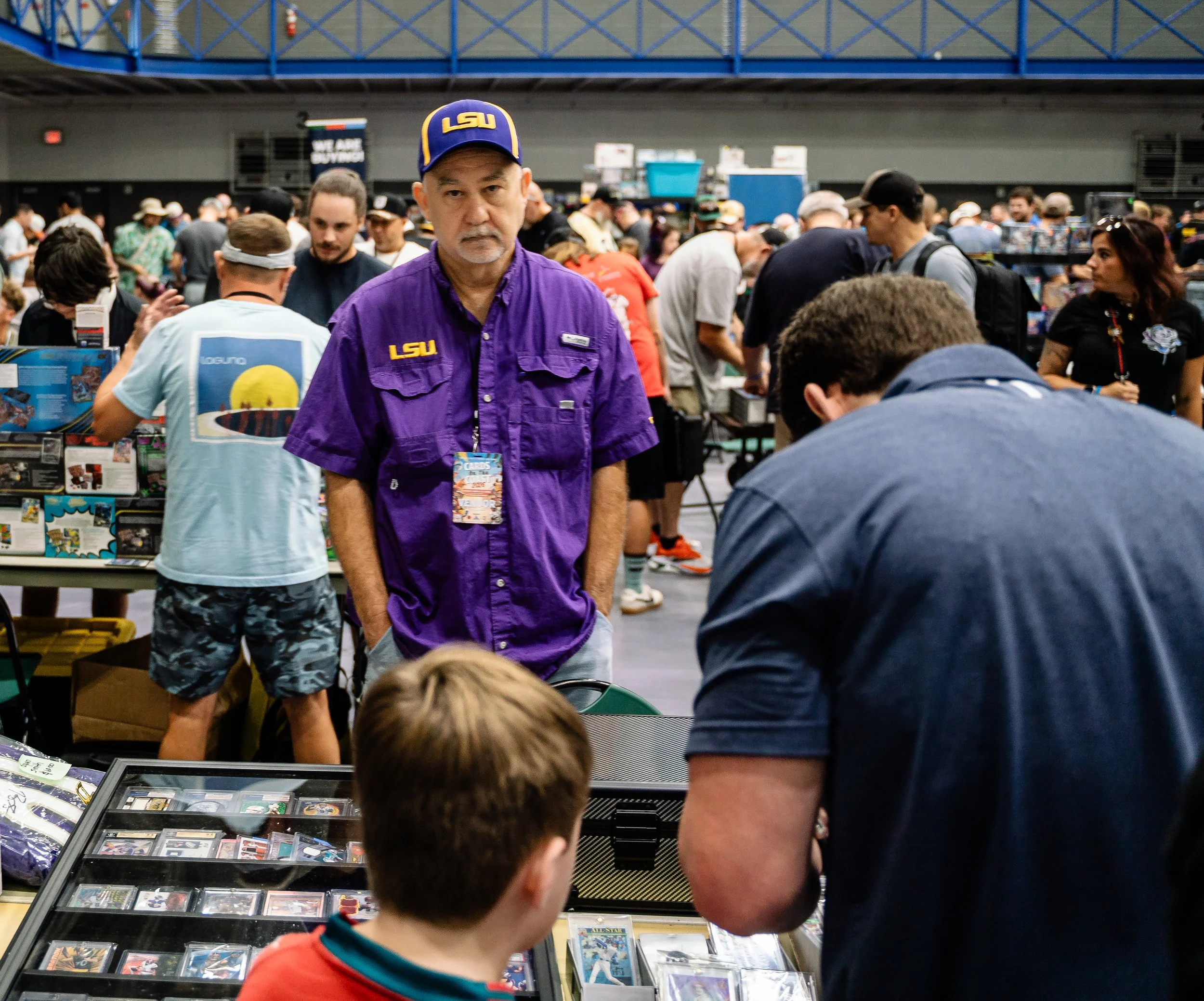 A man wearing a purple LSU shirt and cap stands amidst a crowd at a convention or event, with tables displaying collectibles or cards in front of him.