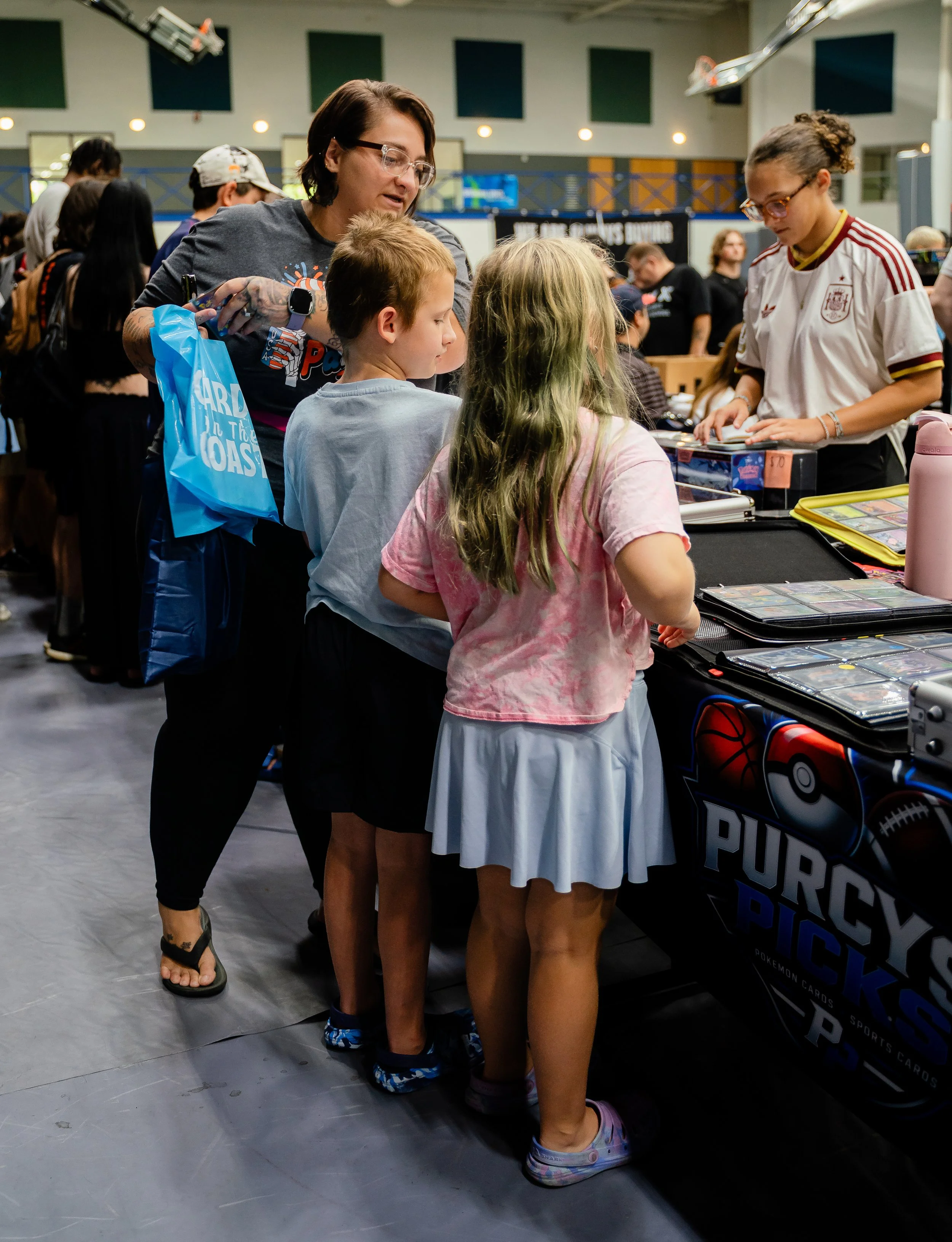 People shopping for Pokémon cards at a convention, with three children and an adult woman gathered around a table displaying Pokémon cards, in a spacious indoor venue.