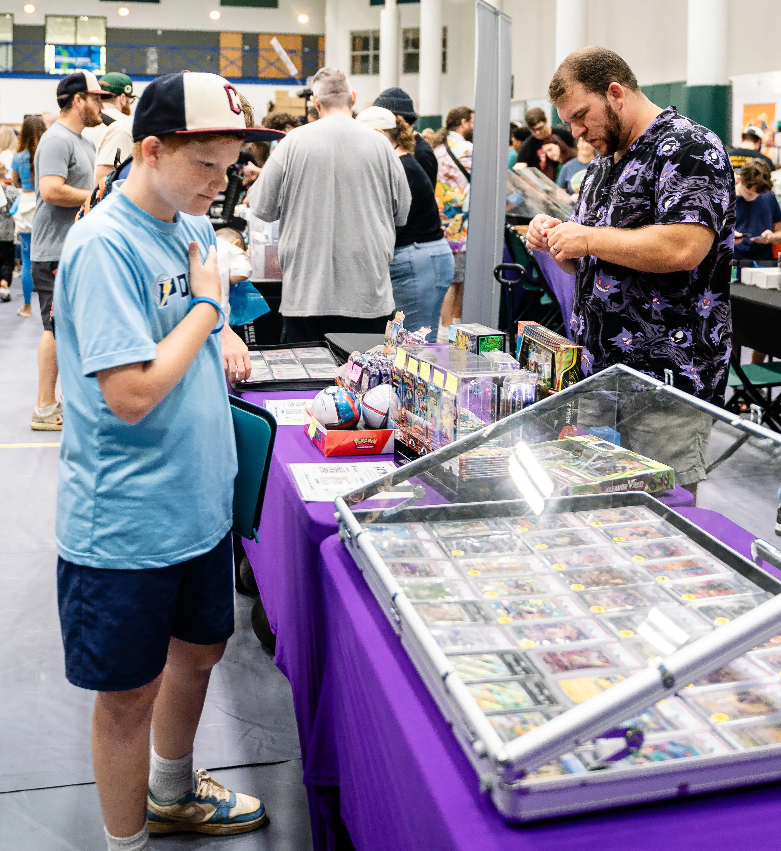 A young boy with a baseball cap and backpack examining Pokémon cards on a display table at a convention or event.