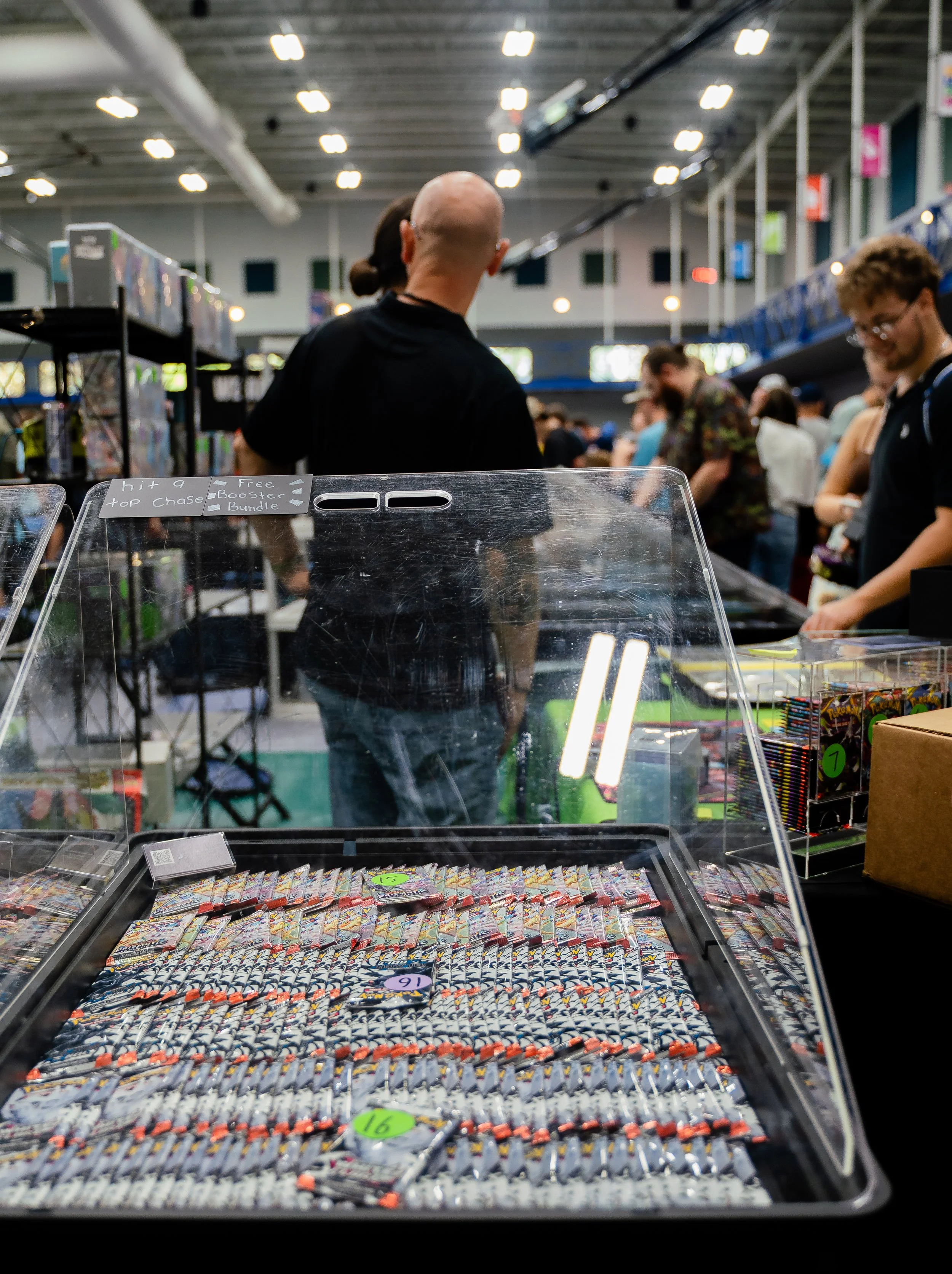 View of a store with people shopping; in the foreground, a display case filled with various candies or small toys, with numbered price tags, and a sign indicating different sales or promotions.