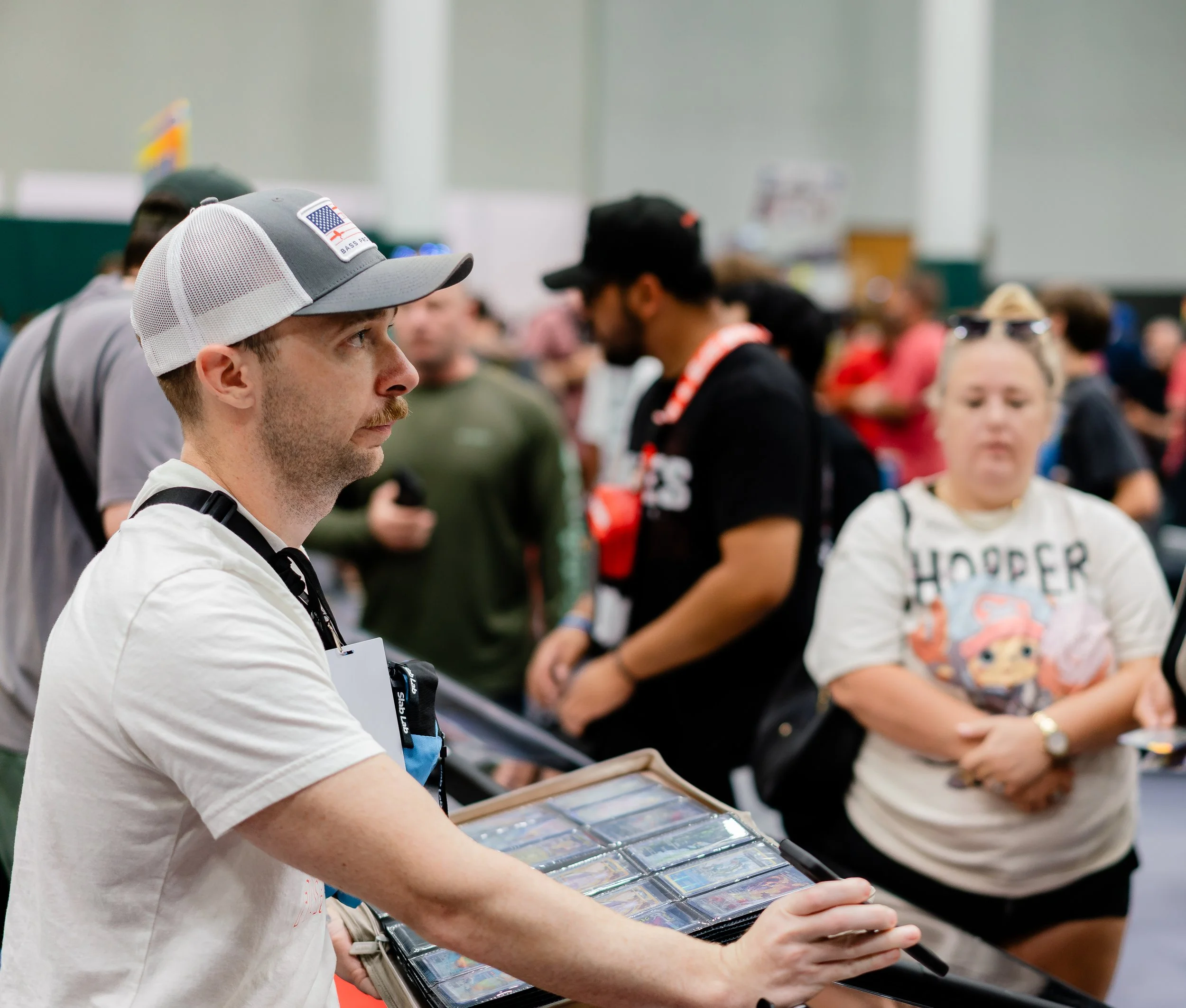A man wearing a white shirt and a gray baseball cap with an American flag patch, looking at a display of trading cards or collectible cards at an indoor event.
