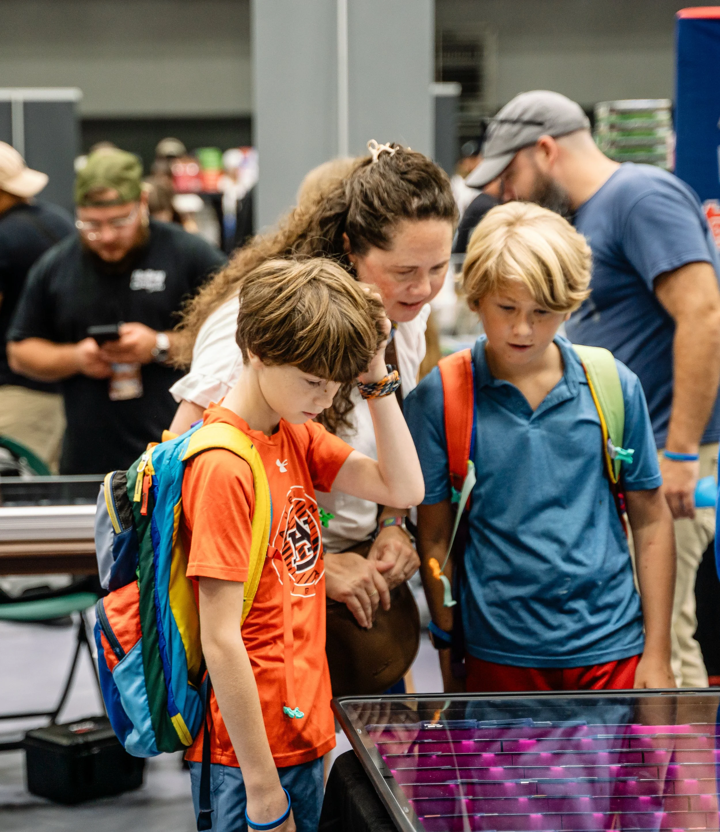 A group of children and adults gathered around an exhibit with purple and pink objects, possibly at a science or educational showcase. The children are wearing backpacks and looking interested.