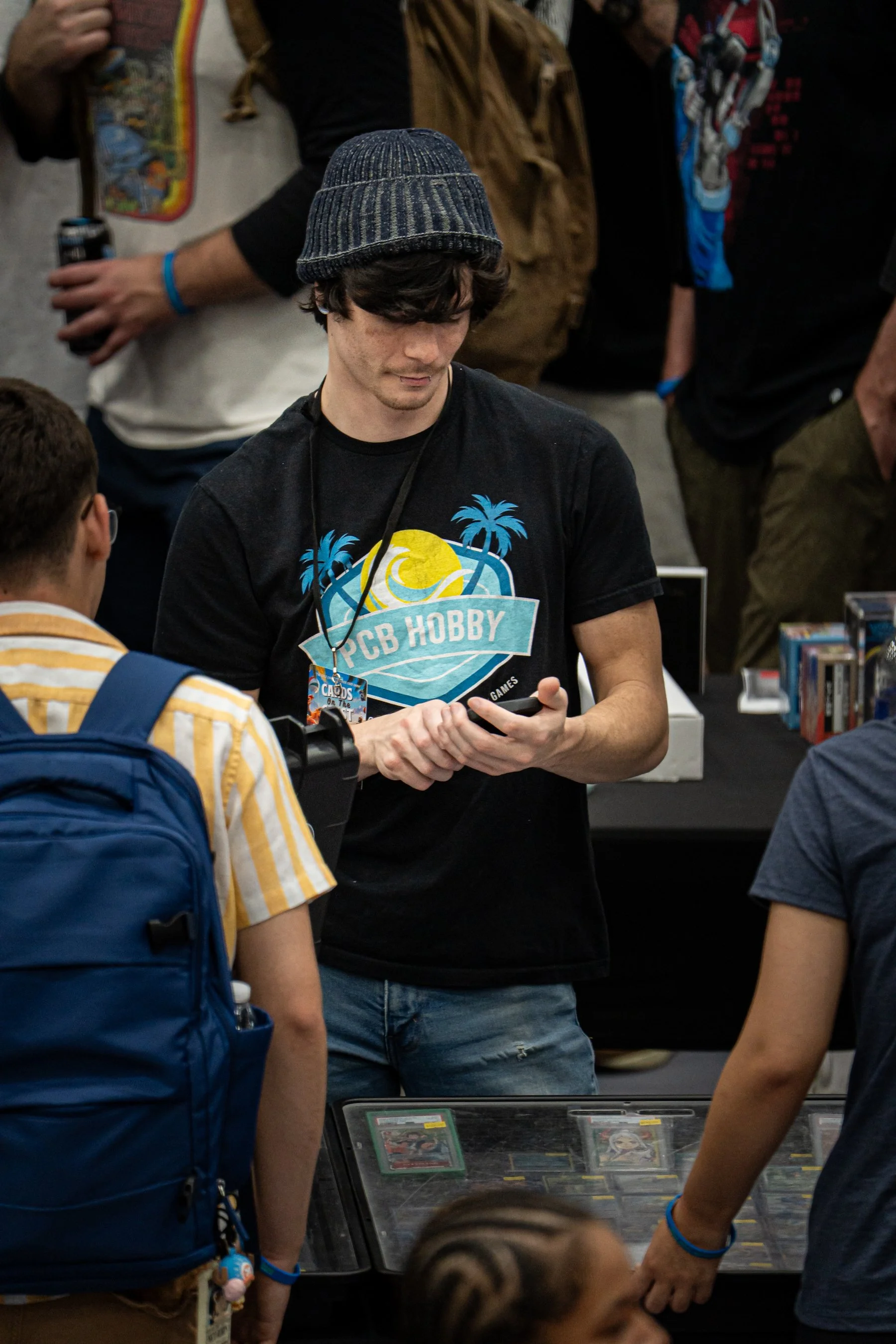 A young man wearing a blue knit beanie and a black T-shirt with a palm trees and sun design is examining trading cards at a convention or store. There are other people around him, with some holding drinks and wearing wristbands.