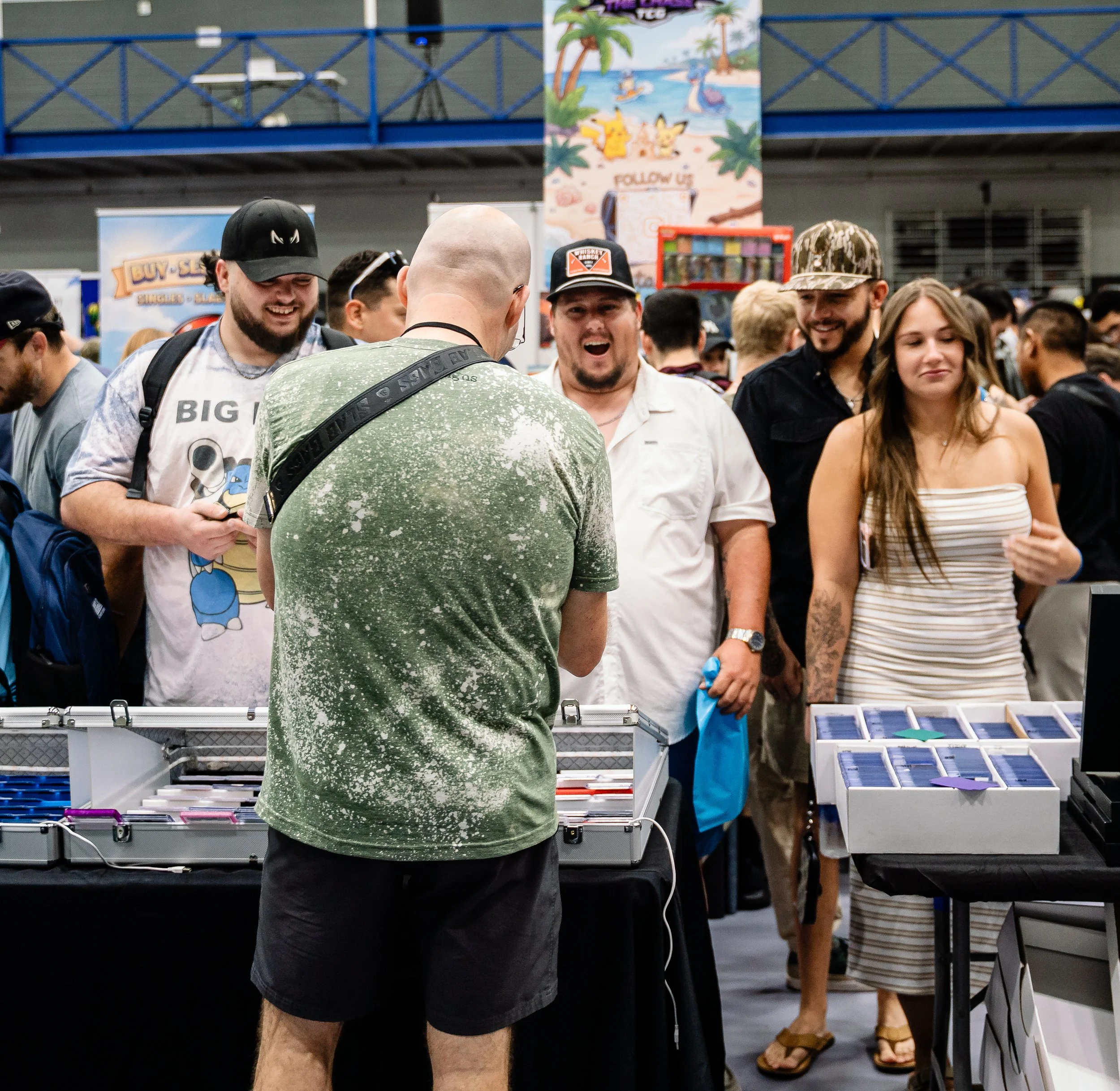 A group of people standing around tables with various electronics and items, at an indoor event or fair. Some individuals are smiling and engaging in conversation, with a colorful tropical-themed poster and other attendees visible in the background.