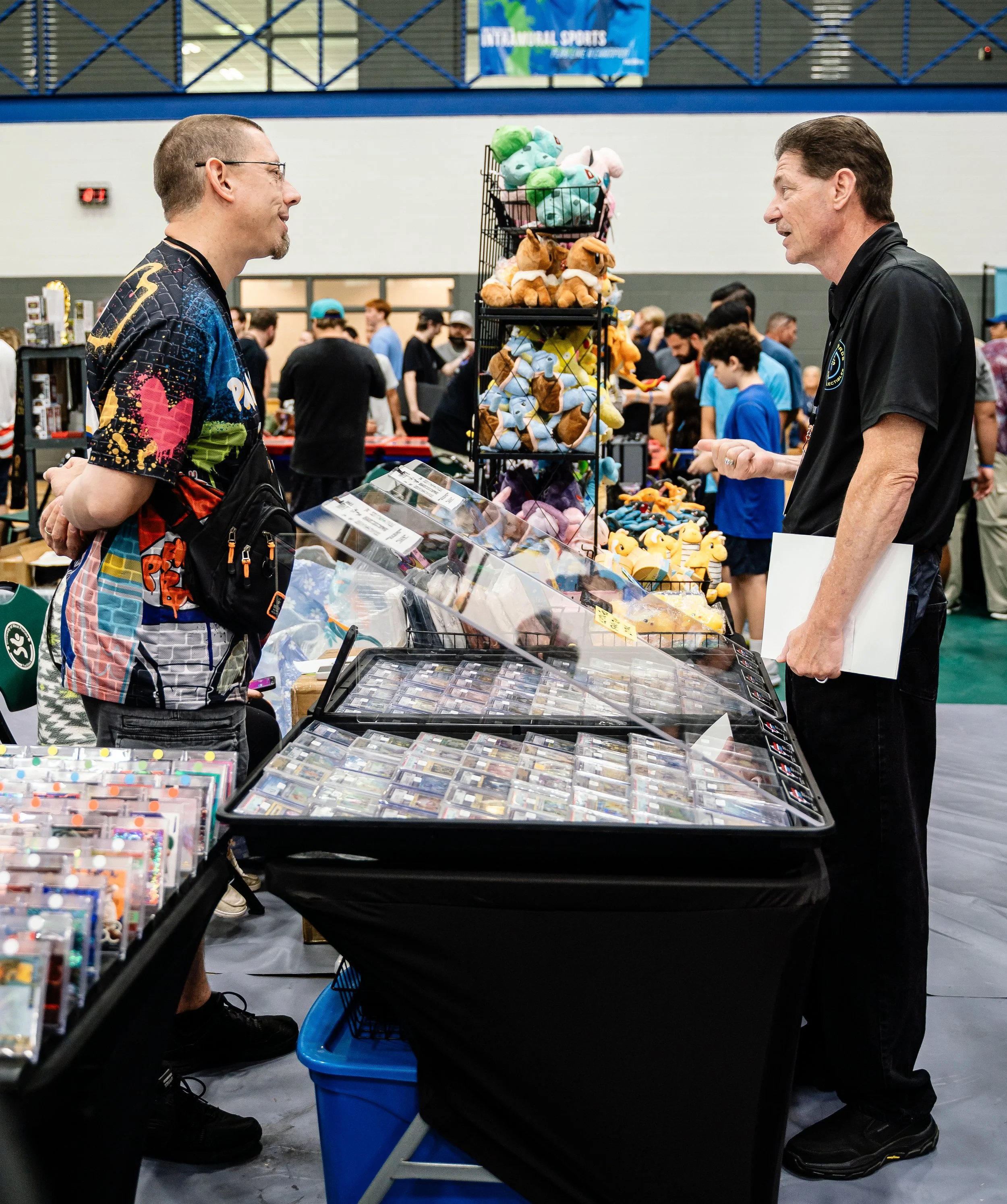 Two men are chatting across a table at a convention or trade show, with various plush toys and collectible items displayed in front of them. The background shows other attendees and booths, indicating an indoor event.
