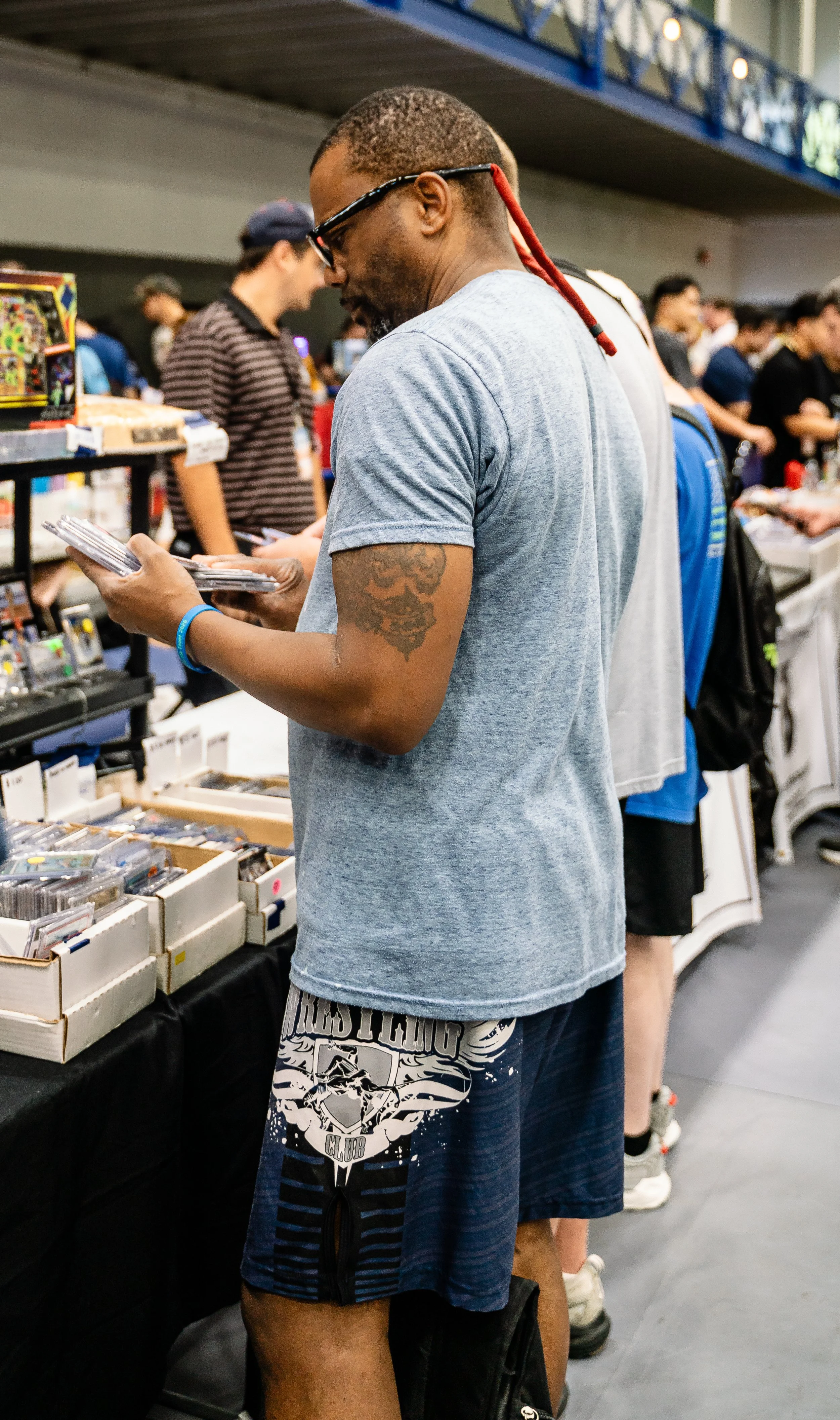 A man wearing glasses and a gray t-shirt is looking at a collection of trading cards at a convention or store. He has a skull tattoo on his upper left arm and is wearing a blue wristband. Other people are visible in the background browsing tables of 