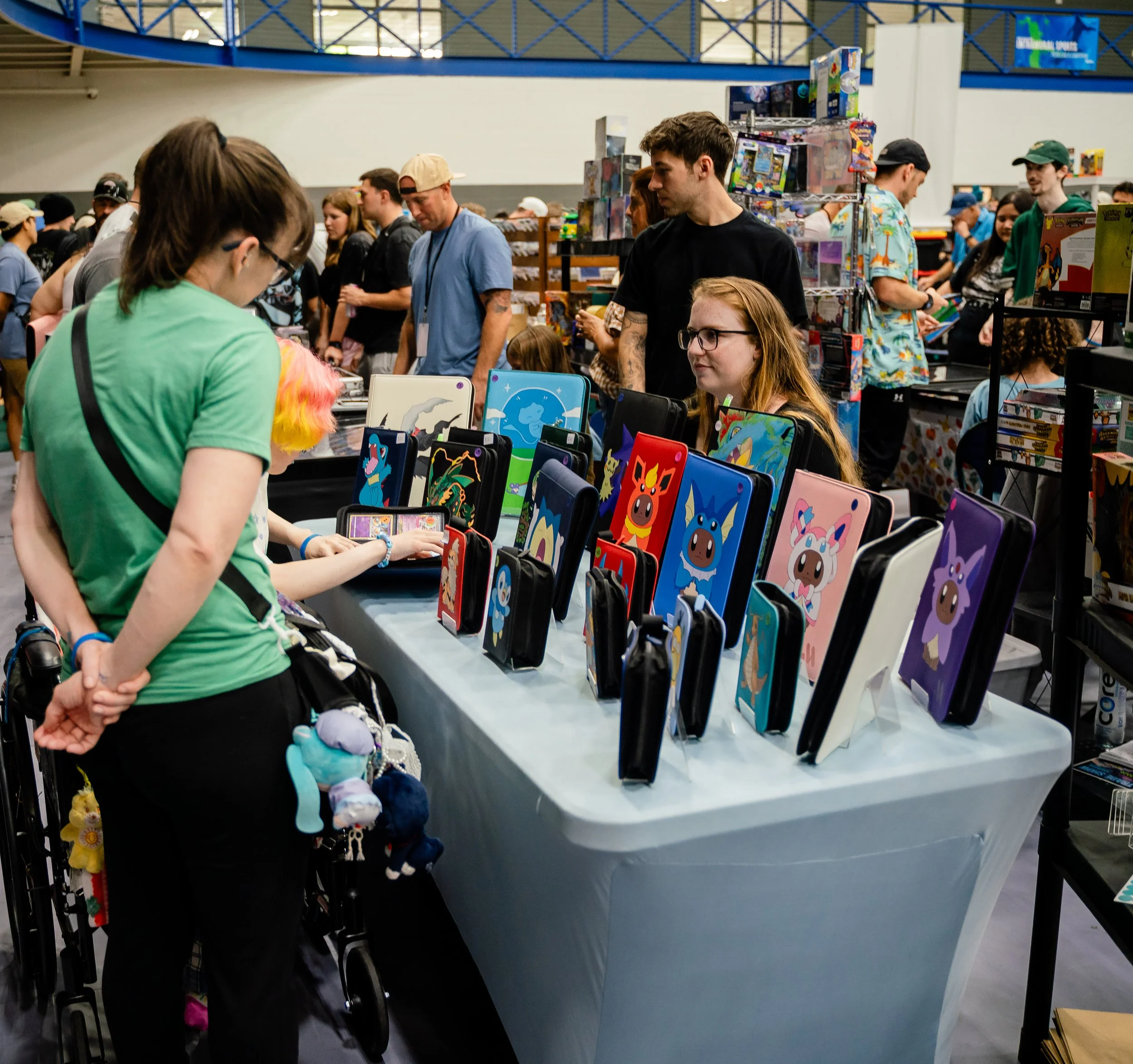 Convention or gaming event with people browsing booths and tables featuring colorful merchandise, pop culture items, and collectibles.