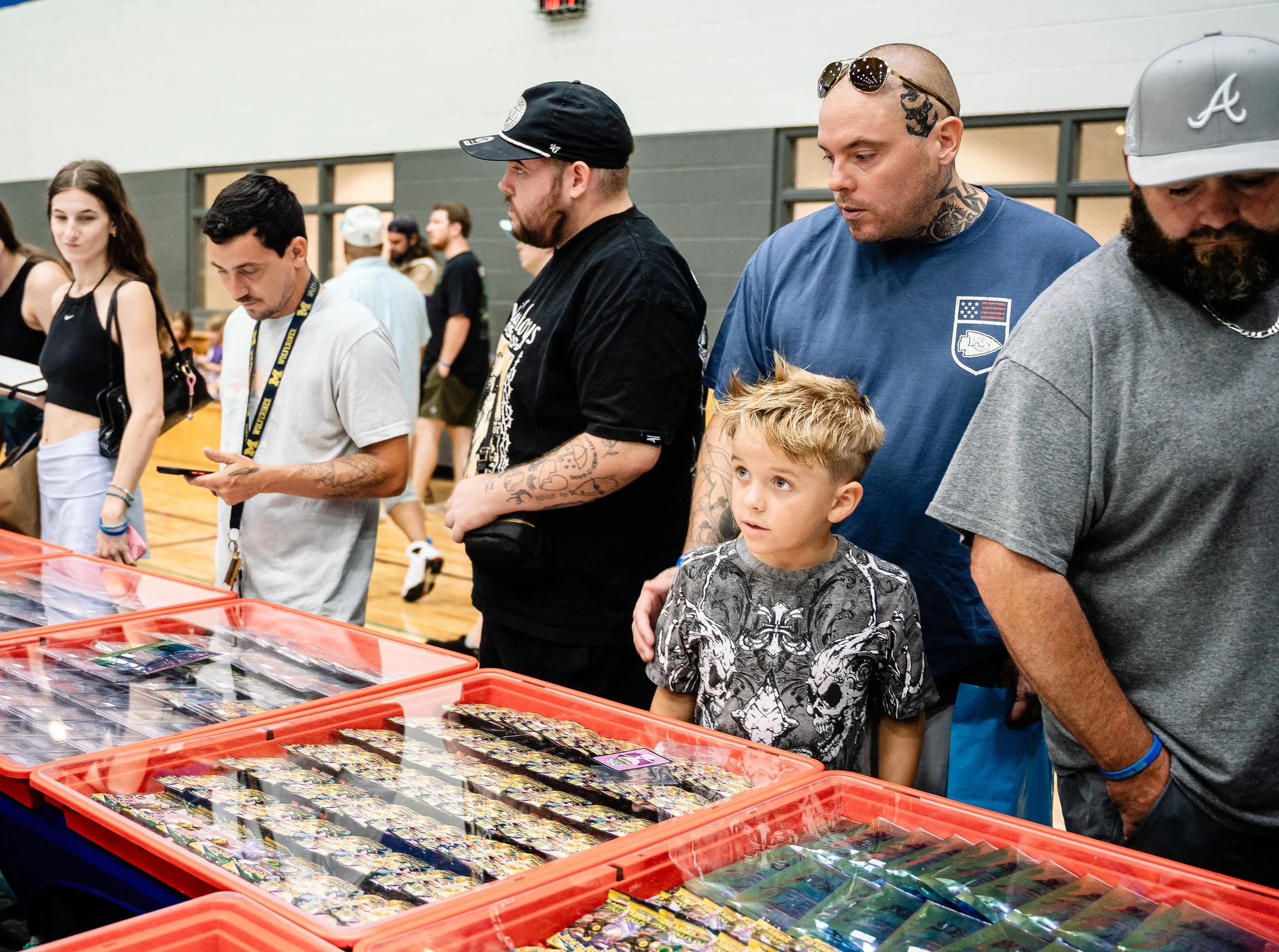 People browsing trading cards at a convention, four men and one boy in the foreground, tables with trading cards in plastic containers.
