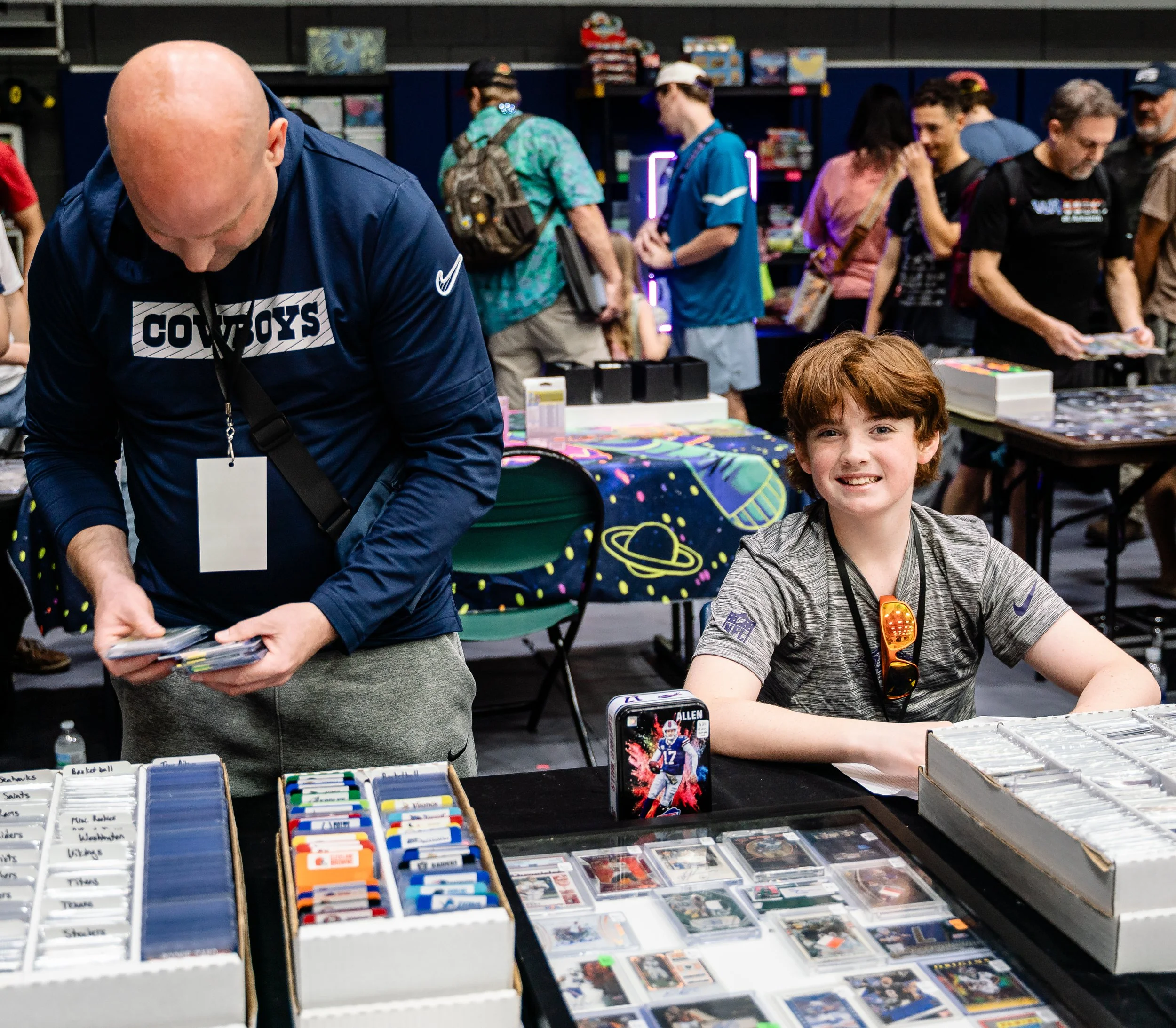 A young boy with brown hair sitting at a table with trading cards, smiling at the camera. Behind him, a man wearing a Dallas Cowboys shirt browses through cards at a stall with organized trading card displays. In the background, there are several peo
