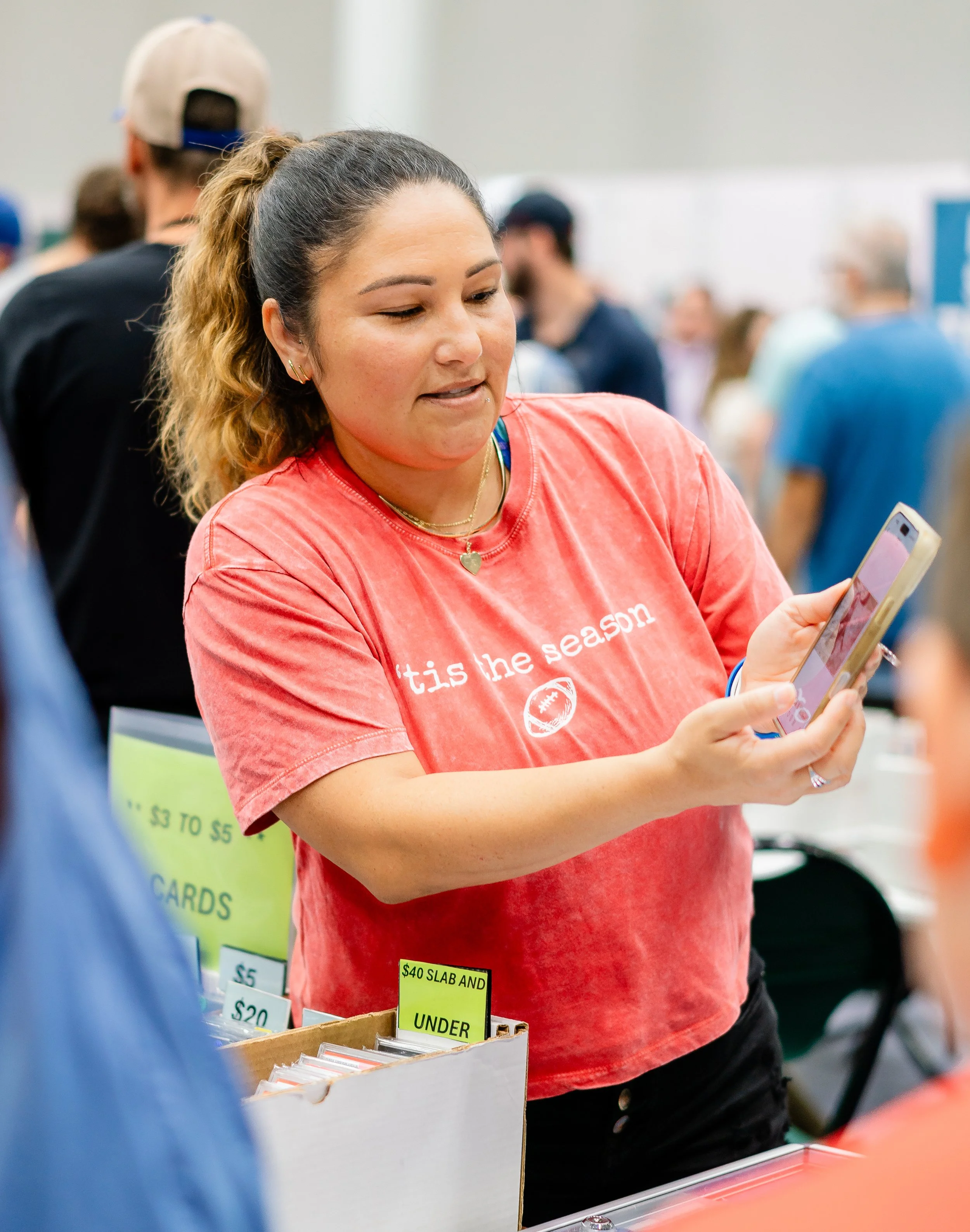 A woman with brown hair in a ponytail wearing a red T-shirt and gold jewelry is looking at her phone at a sale or market booth. The booth has labels with prices such as $40 and $3 to $5, with one label partially visible reading '$40 SLAB AND UNDER.' 