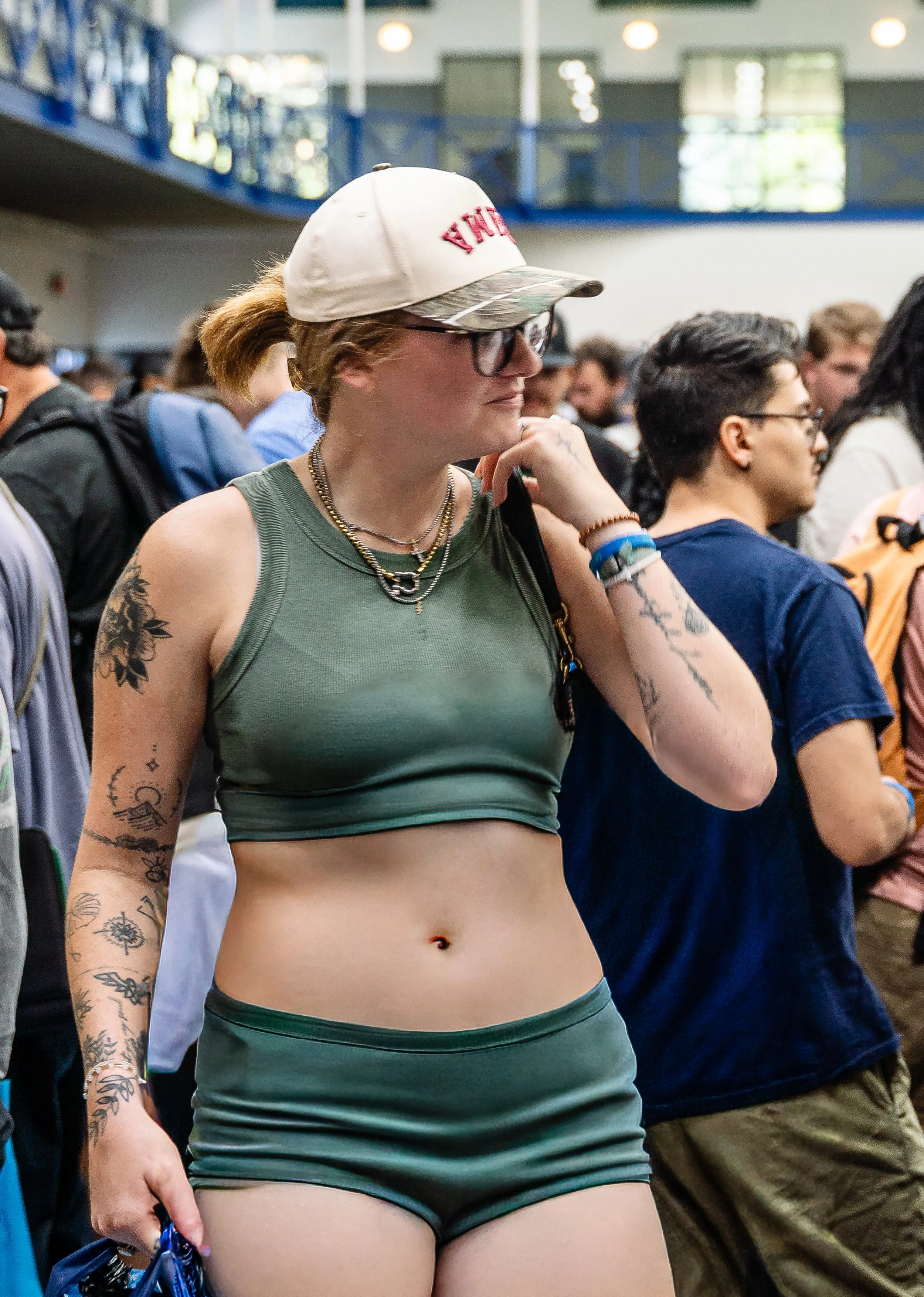 A woman with multiple tattoos wearing a green sports bra and matching shorts, with a white cap and glasses, standing among a crowd indoors.