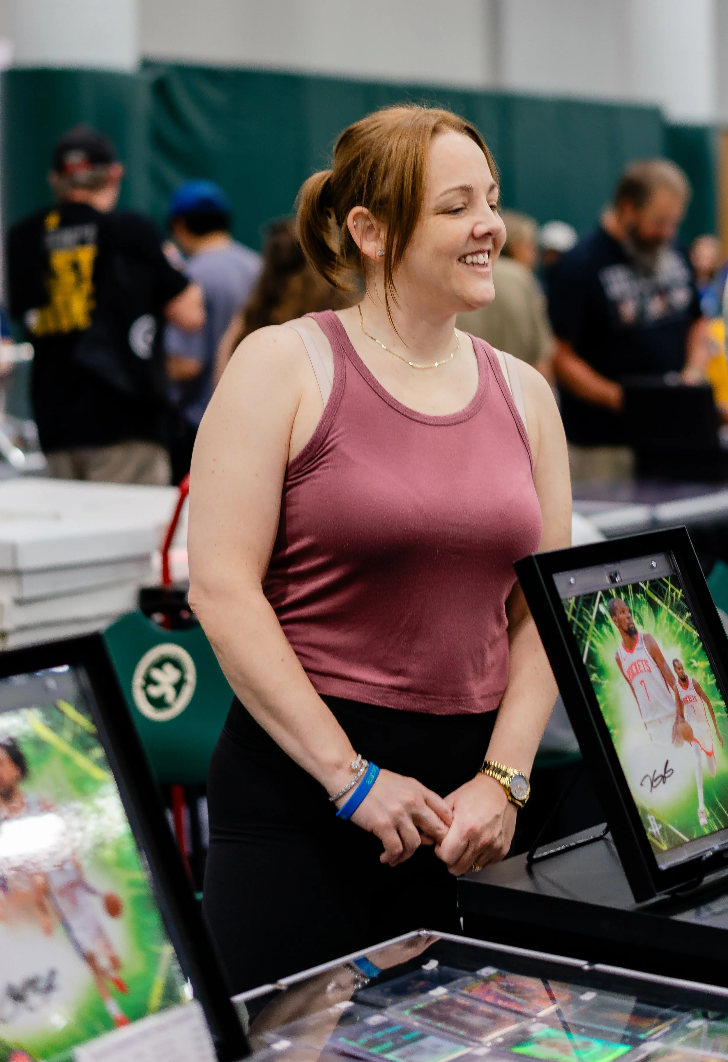 A woman with red hair, wearing a pink tank top and jewelry, standing at a booth with sports memorabilia and electronics, smiling.