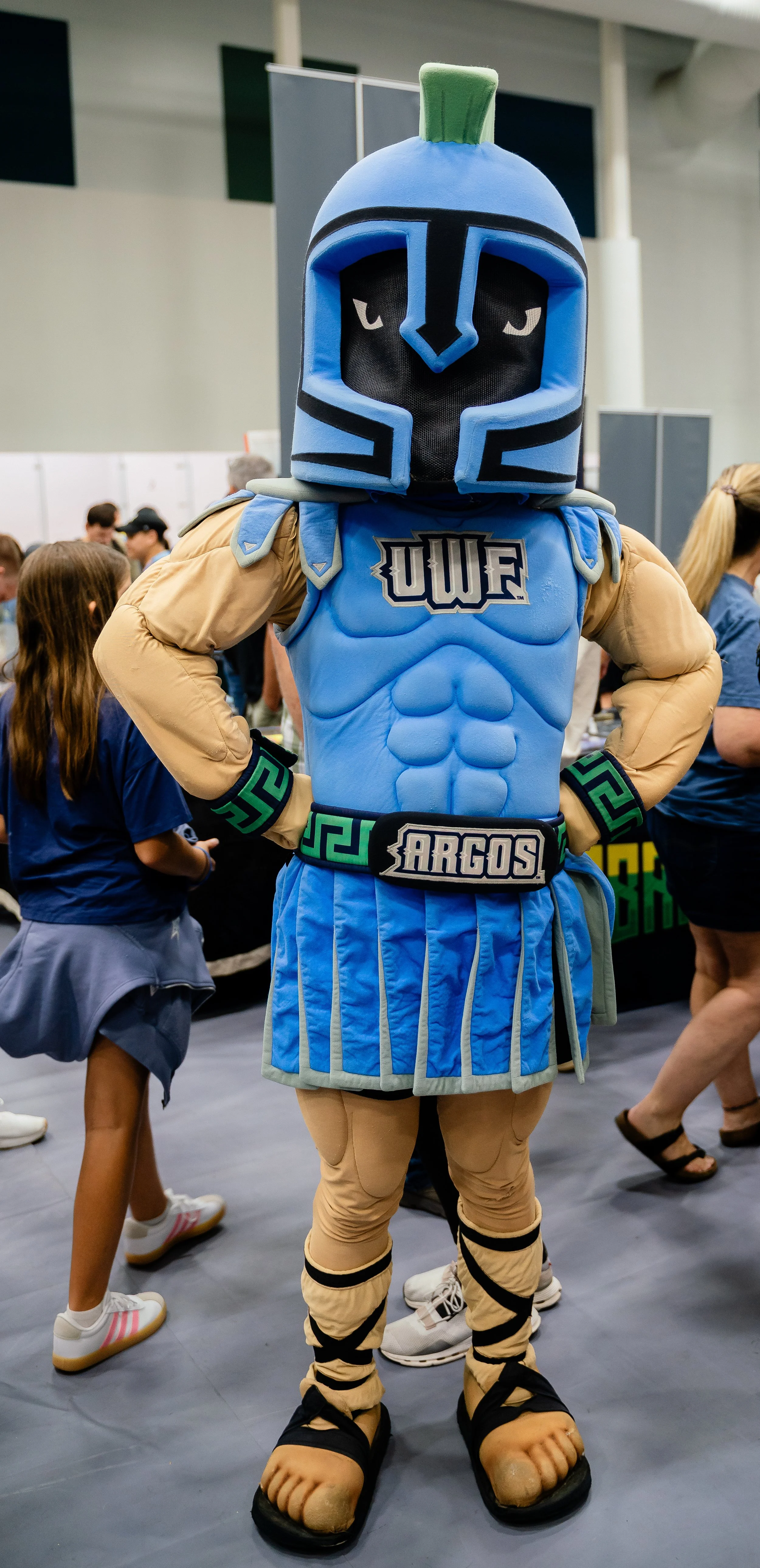Costumed mascot dressed as a Greek soldier with a helmet, blue armor, and sandals, standing with hands on hips in a crowded indoor event.
