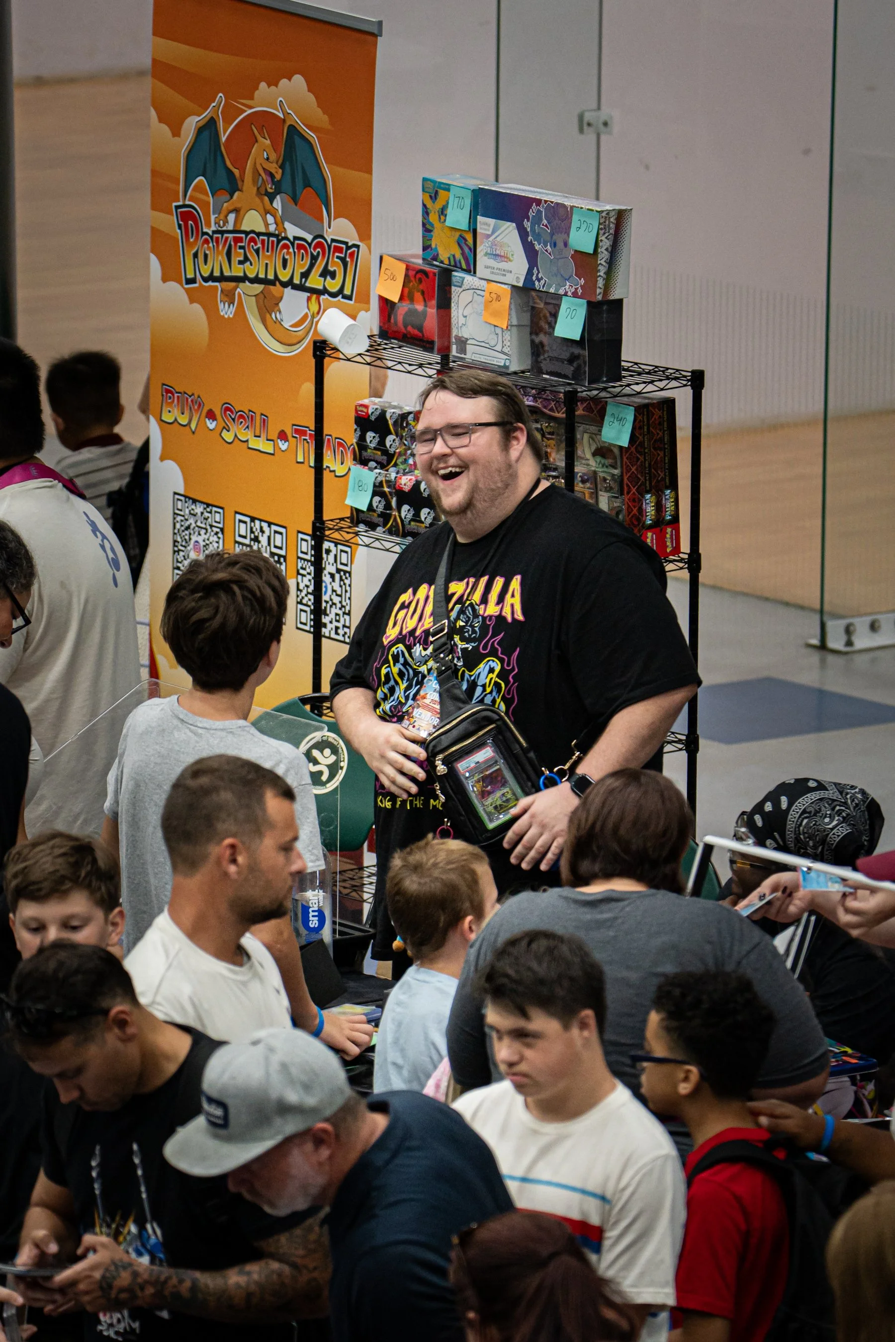 A crowded indoor event with a man wearing a black T-shirt featuring Marvel's Black Panther, smiling and talking to children and other people. Behind him is a sign for Pokeshop251 with a Pokémon logo, boxes of trading cards, and QR codes.