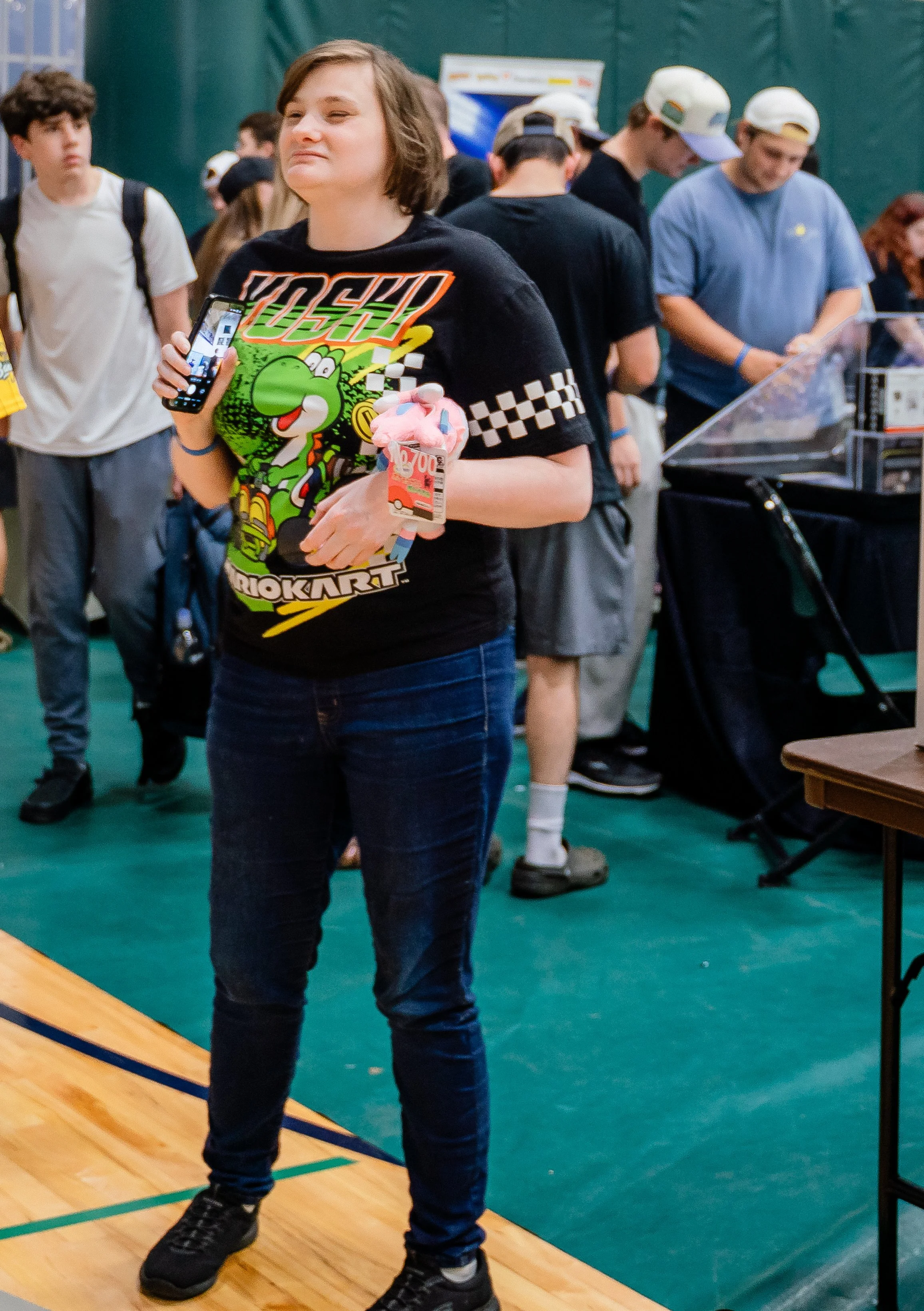 A young woman standing on a wooden basketball court with a green floor, holding a phone and a plush toy, with a group of teenagers in the background at an indoor event.