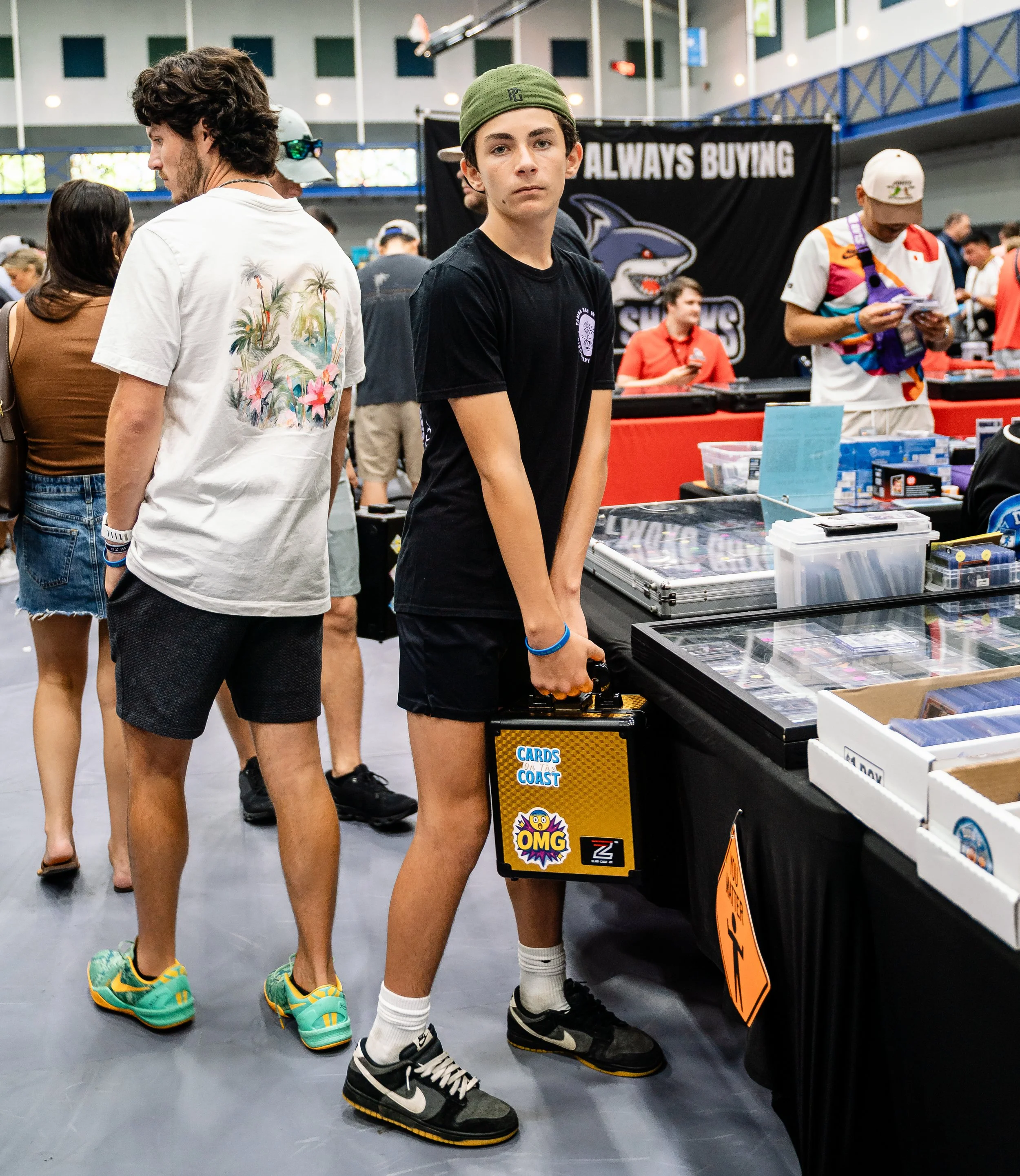 Two young men at a convention or event, standing next to tables with collectibles or trading cards. The one in the foreground has dark hair, is wearing a black T-shirt, a green cap, and black Nike sneakers with white socks. The other in the backgroun