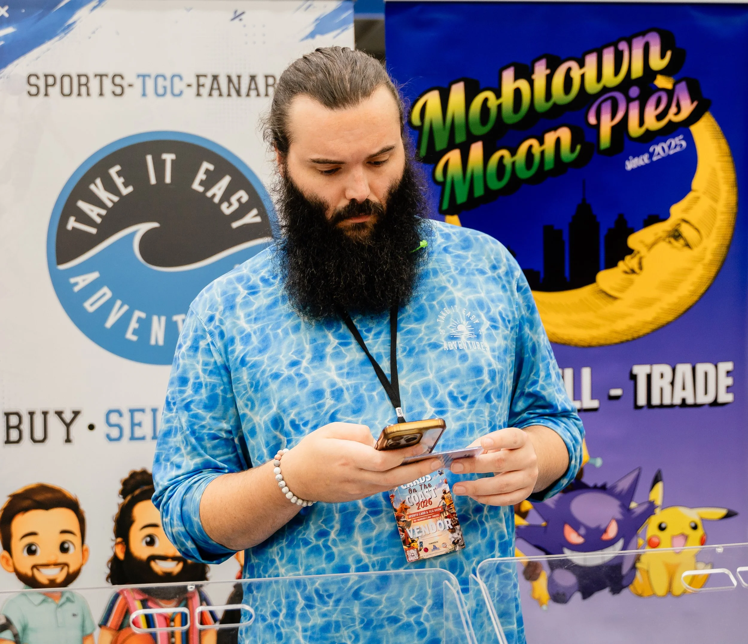 A man with long hair and a beard looking at his phone at an event with banners and cartoon characters in the background.