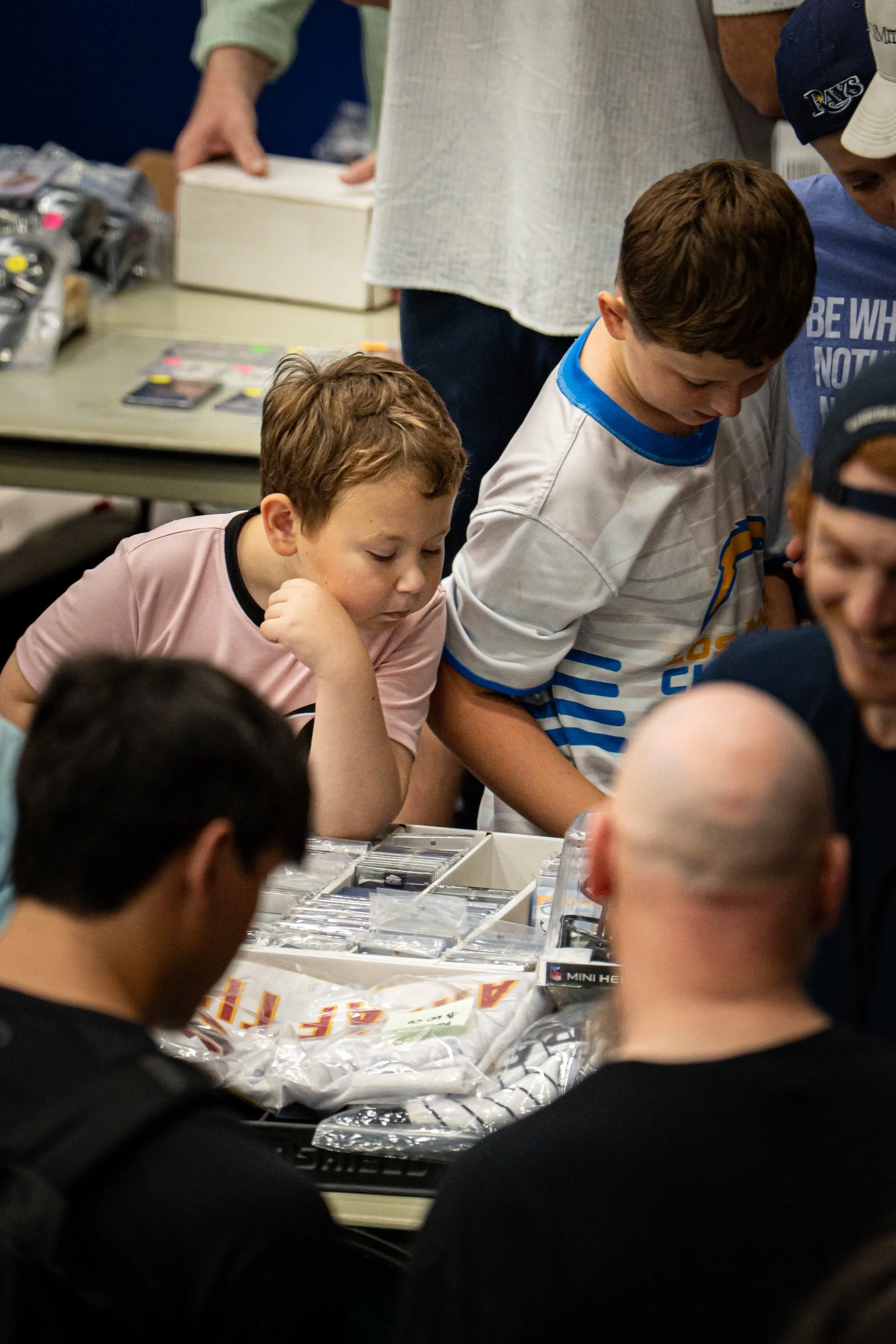A group of children and adults gathered around a table, looking at items arranged in plastic containers, possibly trading cards or collectibles at an indoor event.