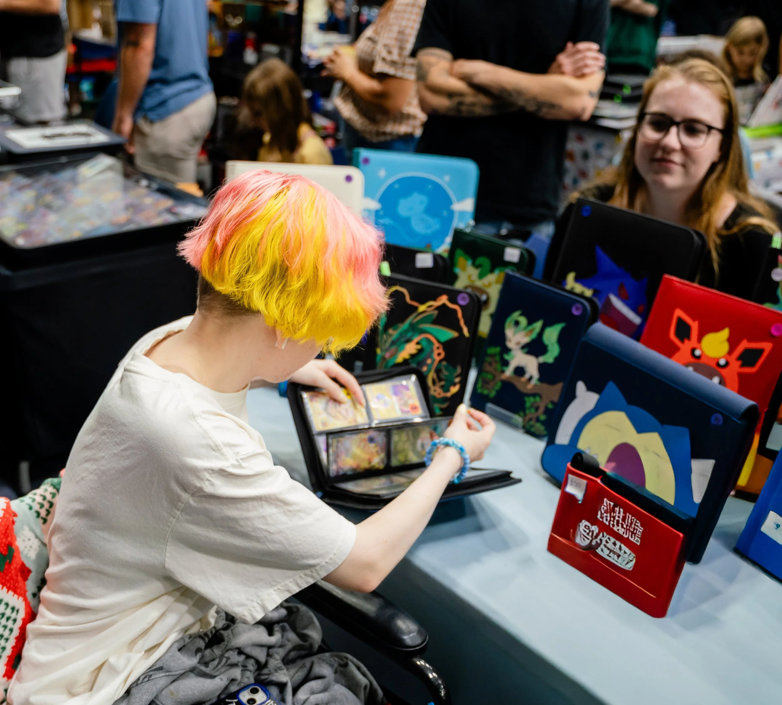 A person with brightly colored hair, primarily yellow and pink, sitting at a table and using a tablet device in a crowded indoor event, possibly a convention or fair, with display booths and other people in the background.