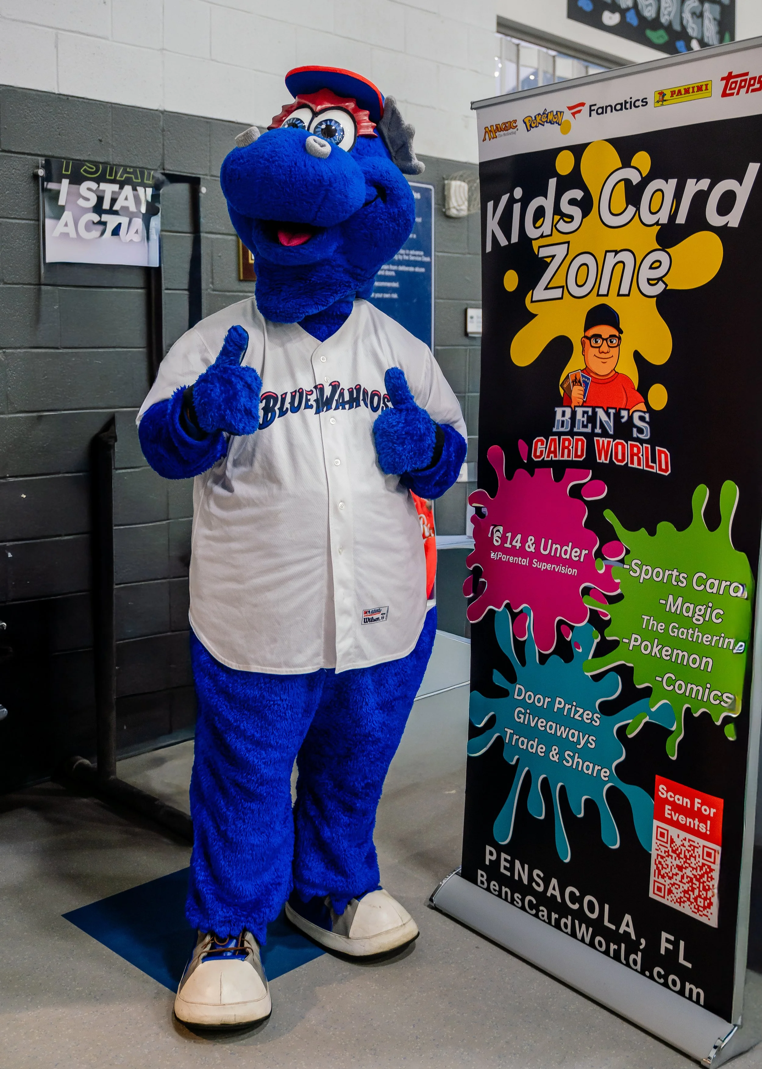 A person in a blue mascot costume resembling a dog wearing a white baseball jersey, red and blue cap, sneakers, and giving two thumbs up, standing next to a sign for Ben's Card World in Pensacola, Florida.