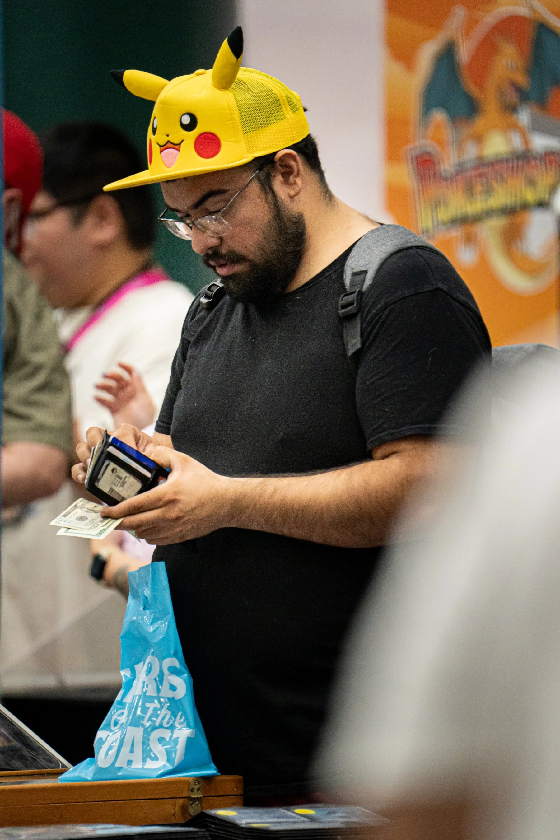 A man wearing glasses, a Pikachu hat, and a black t-shirt, holding cash and a wallet at a convention or event.