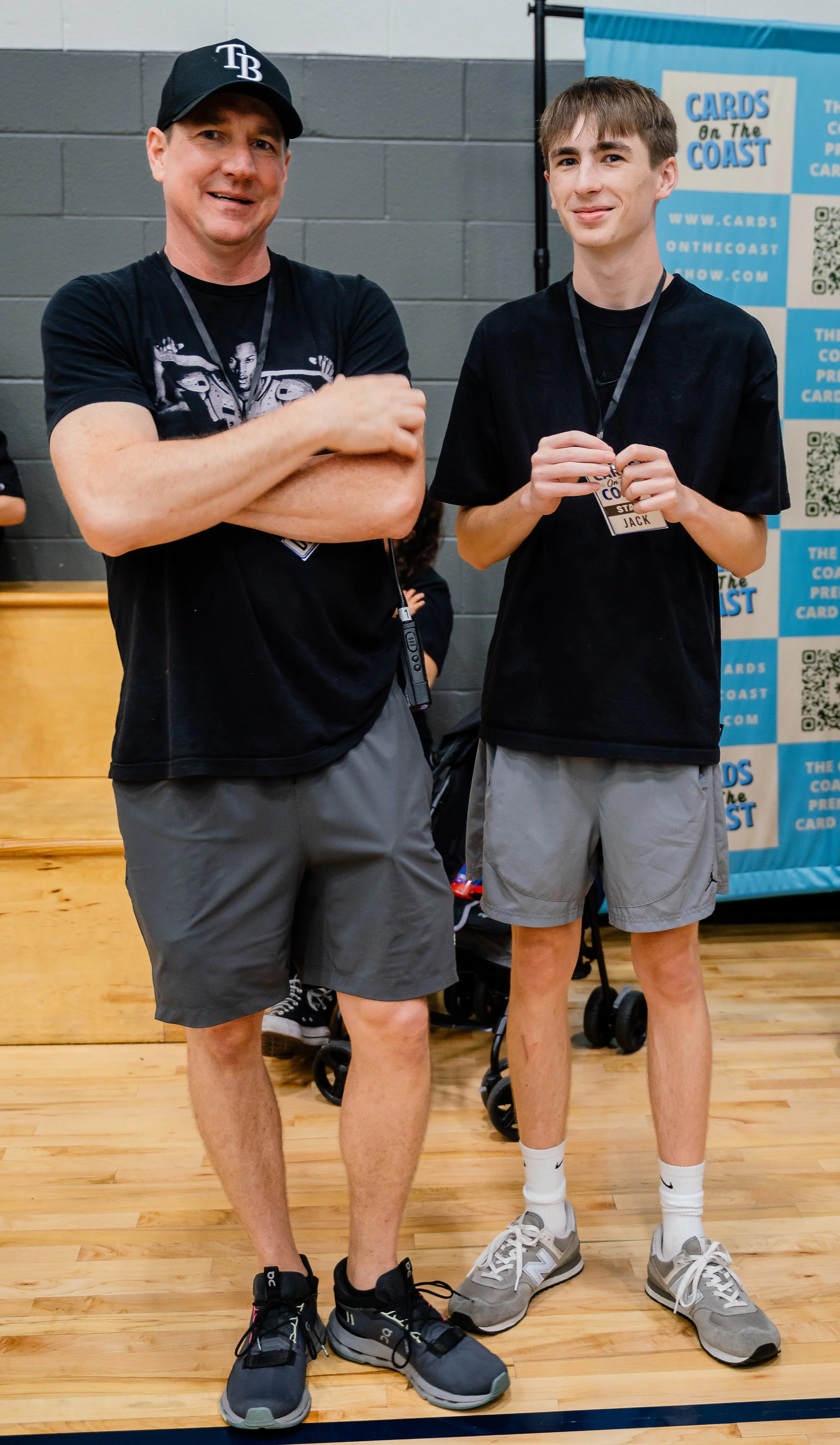 Two young men standing indoors on a wooden floor, wearing black t-shirts and gray shorts, with one wearing a baseball cap and the other holding a name tag, and a banner in the background that reads 'Cards on the Coast'.