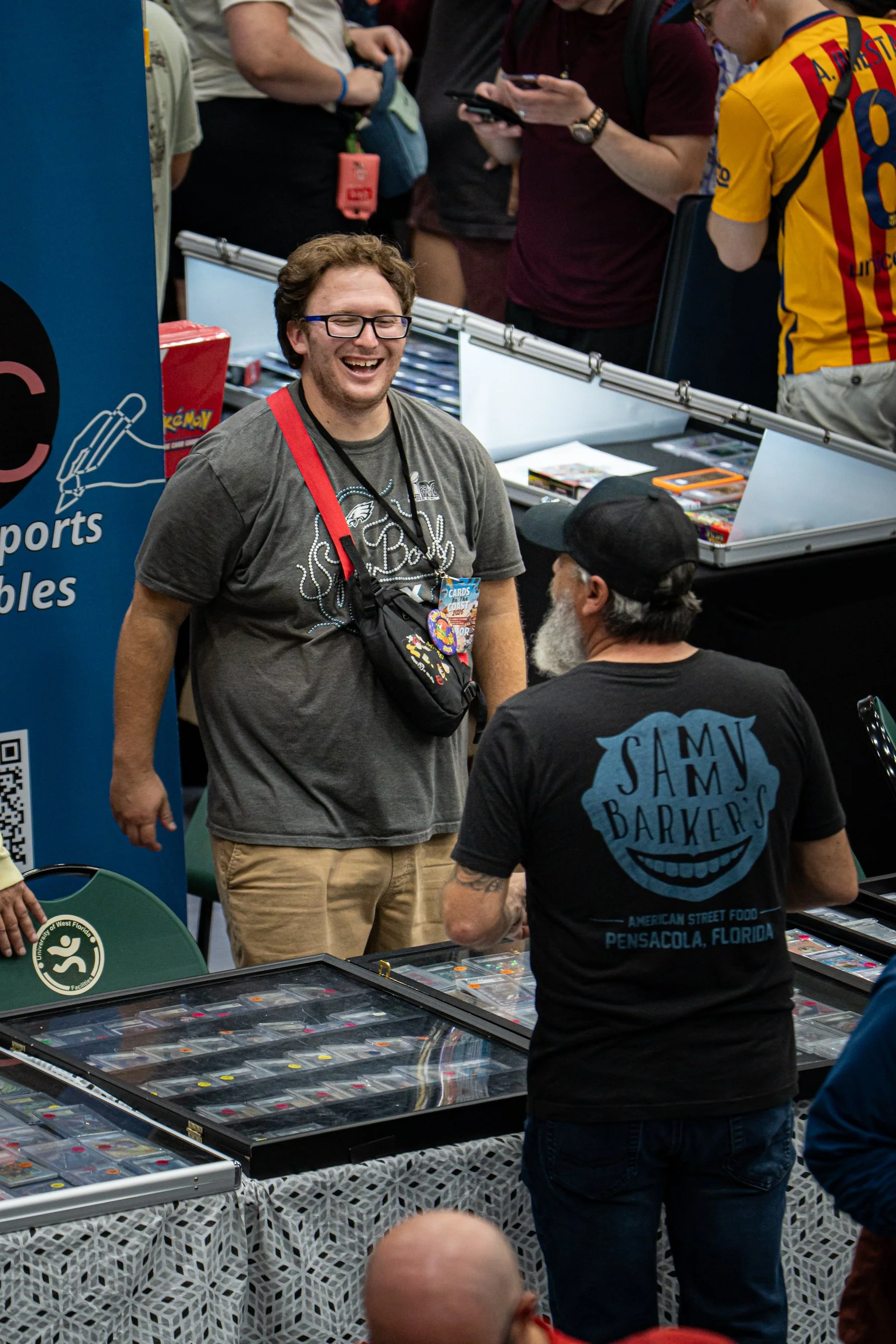 People at a convention or event looking at and talking about displays of toy cars and model kits, with some individuals using smartphones and wearing event badges around their necks.