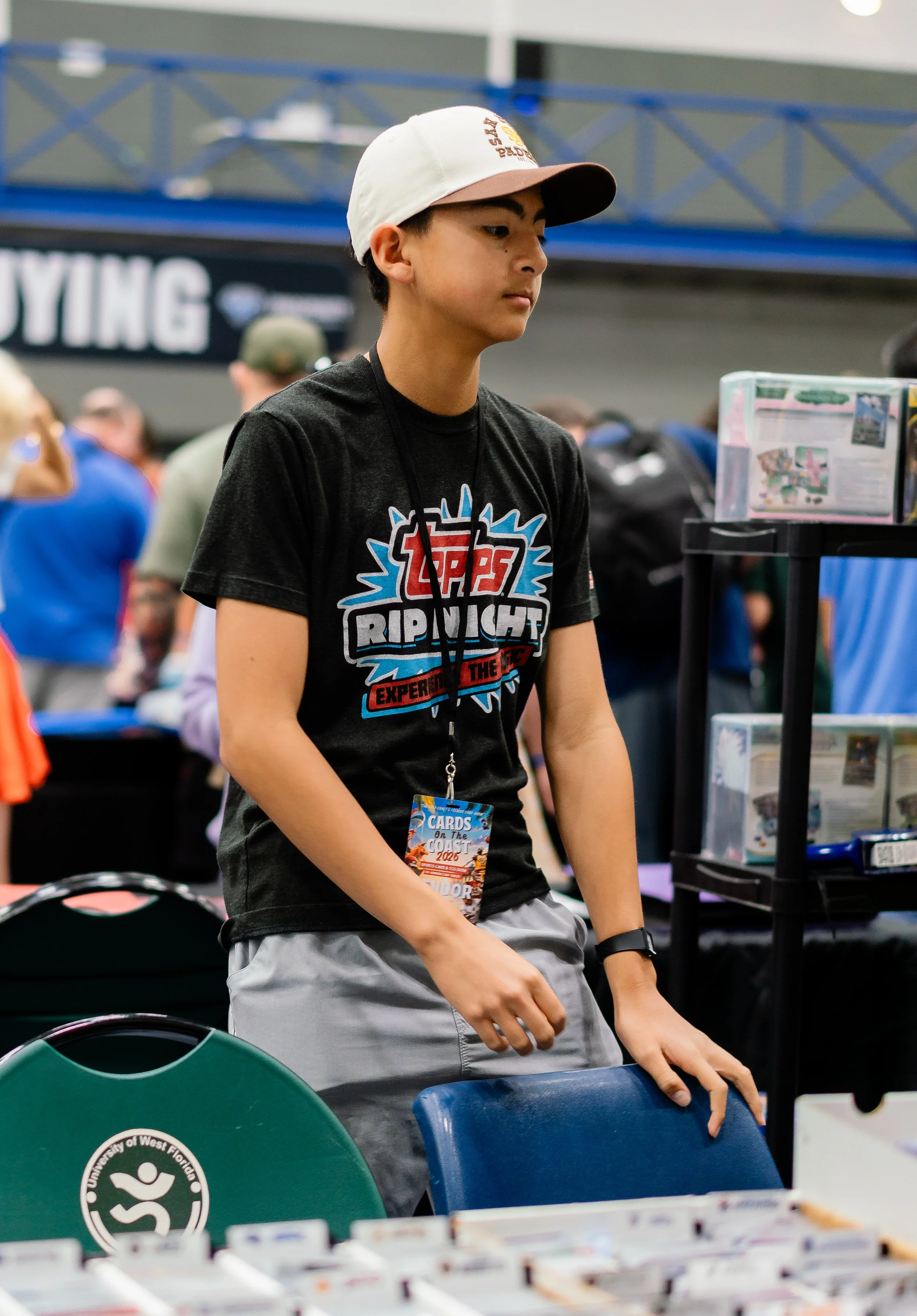 A teenage boy with short dark hair wearing a white and light brown baseball cap, a black T-shirt with colorful graphics, and gray shorts standing at a table at a convention or event. The background shows other people, a sign, and display shelves with