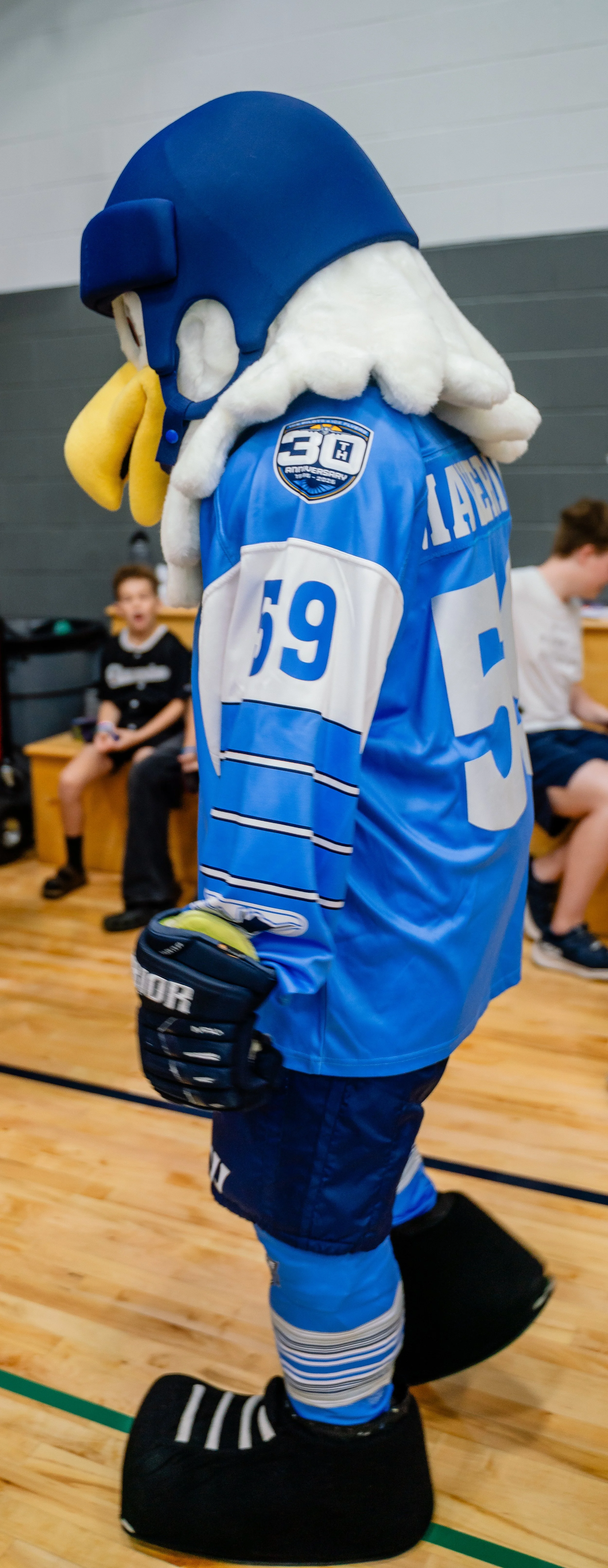 Costumed mascot dressed as a hockey player standing on a padded platform in a gymnasium, with children sitting in the background.