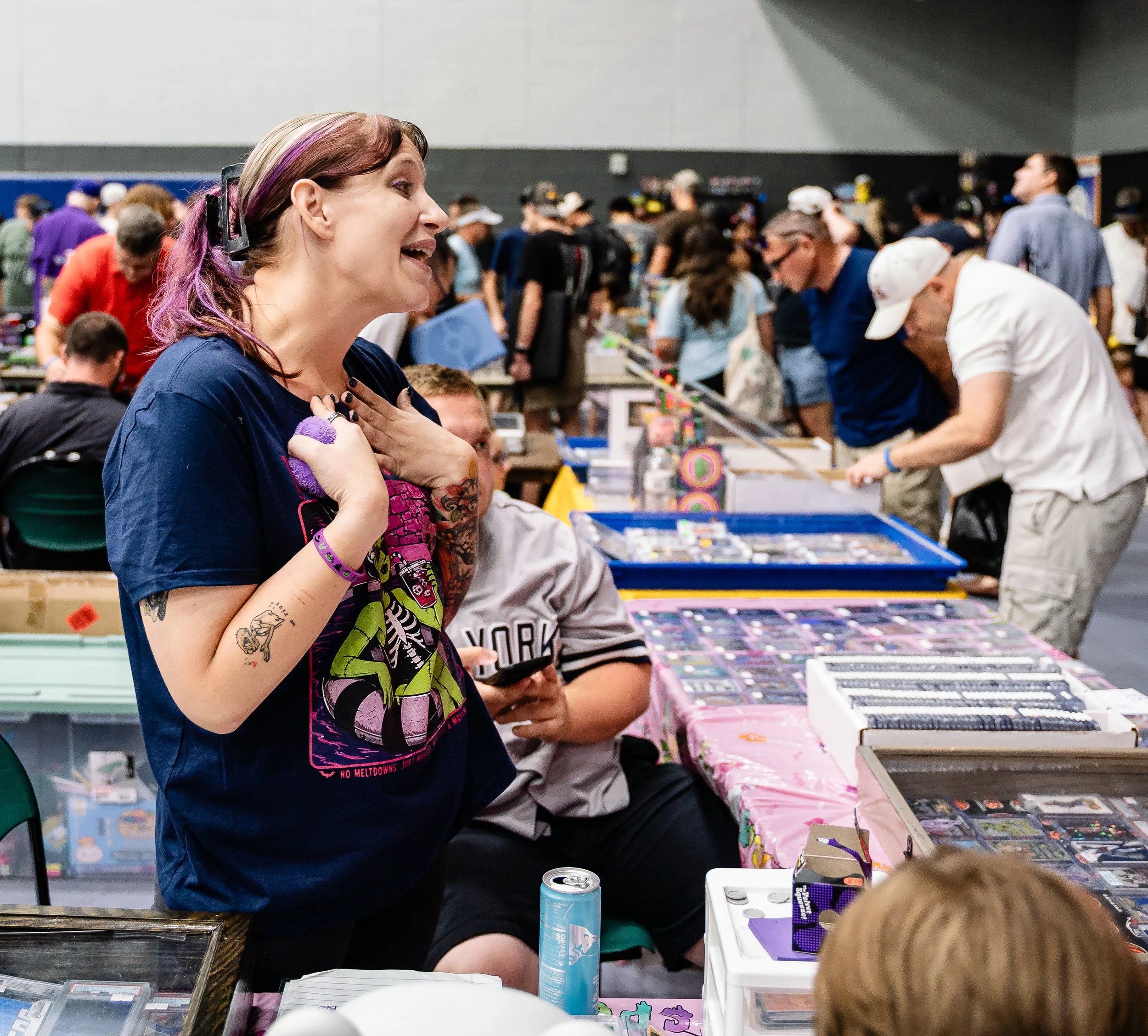Woman with purple hair and tattoos smiling at a crowded indoor event with tables and people browsing collectibles or items.