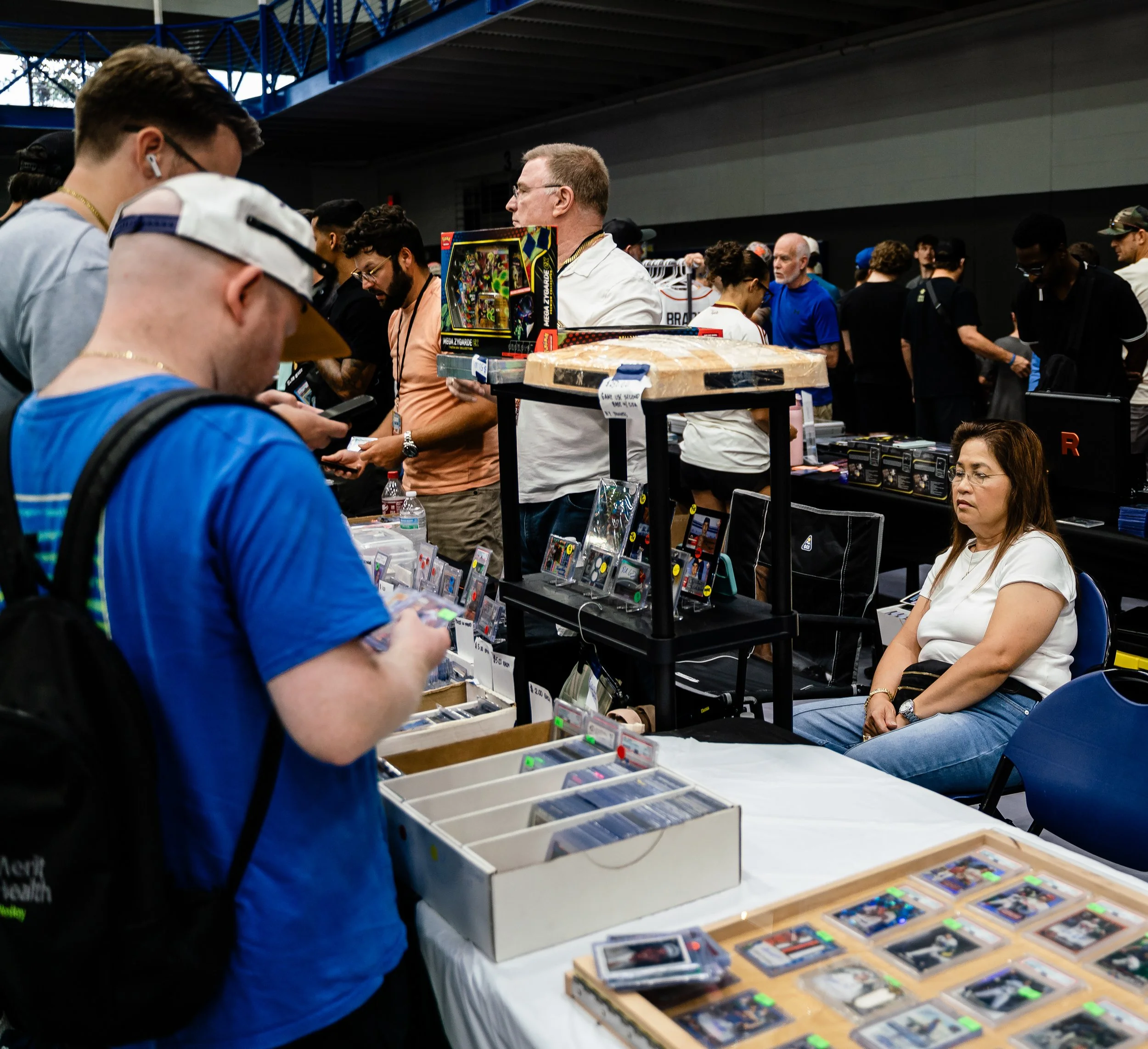 People browsing collectible cards and toys at a convention or market, with tables displaying various merchandise.